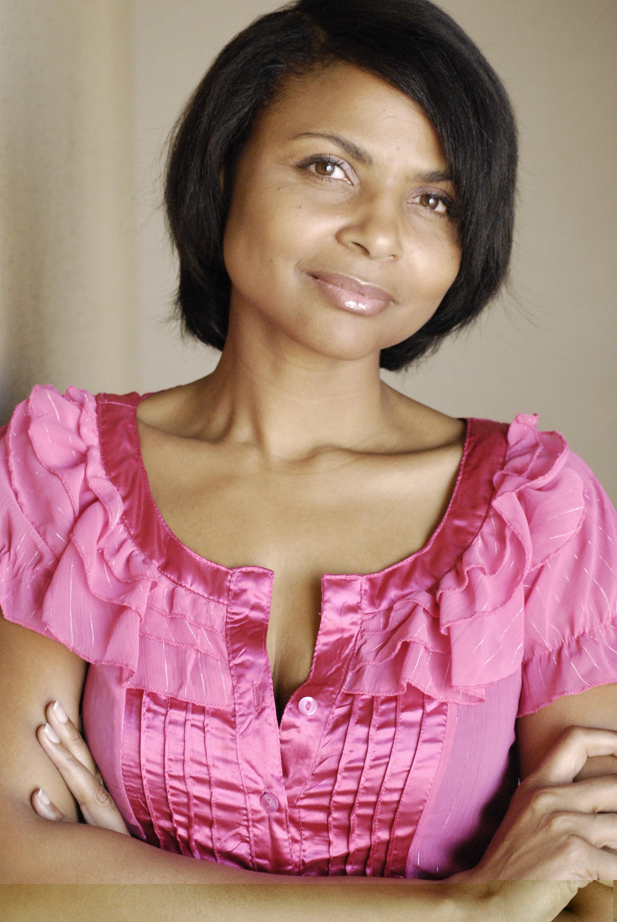 A woman with short black hair, wearing a pink ruffled dress, standing with her arms crossed.
