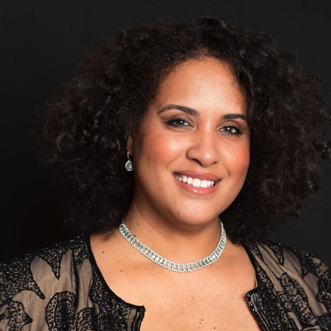 Portrait of a woman with curly dark hair wearing a black beaded dress and diamond jewelry, smiling against a black background.