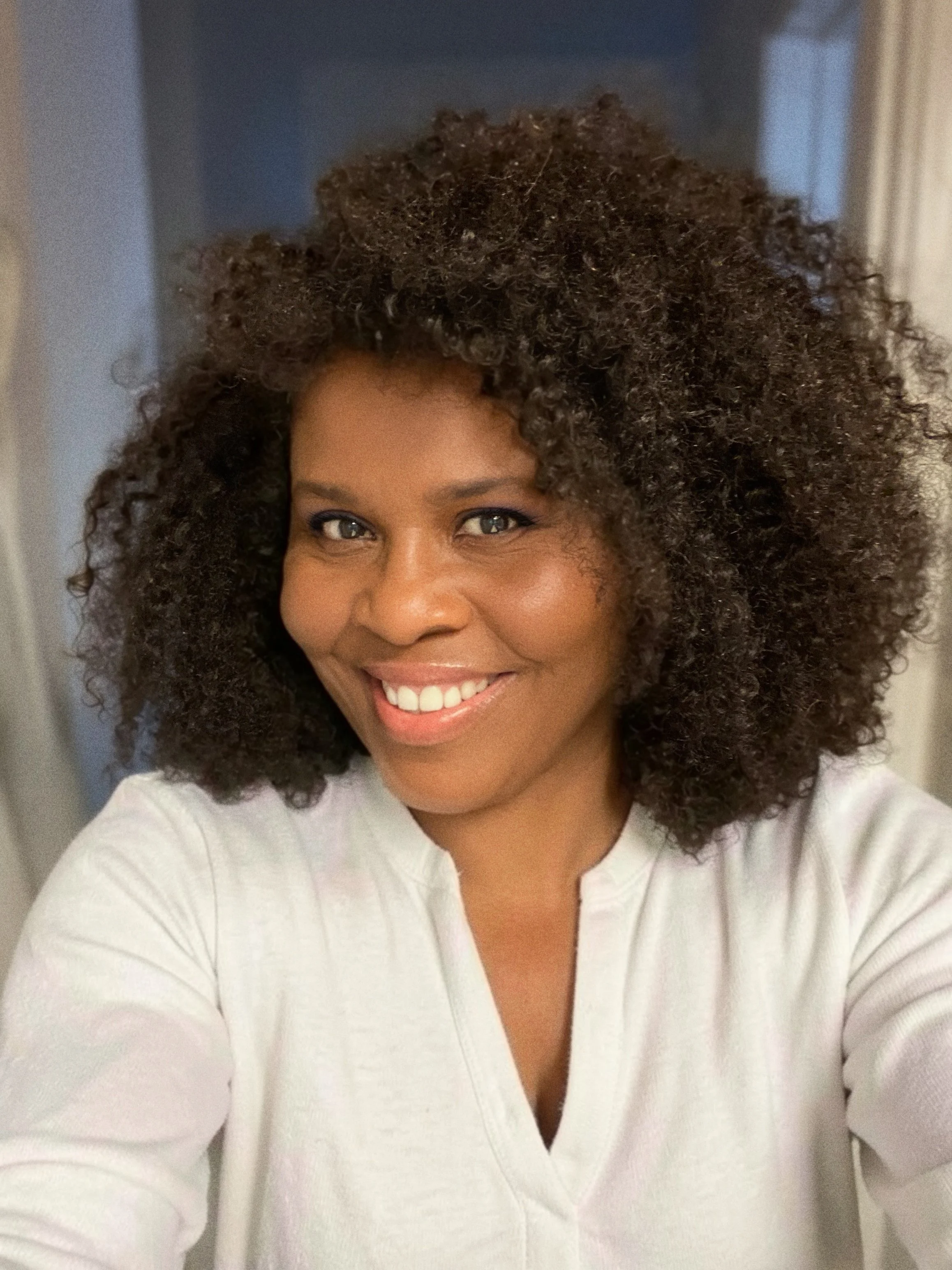 A woman with a wide smile, curly natural hair, and wearing a white top.