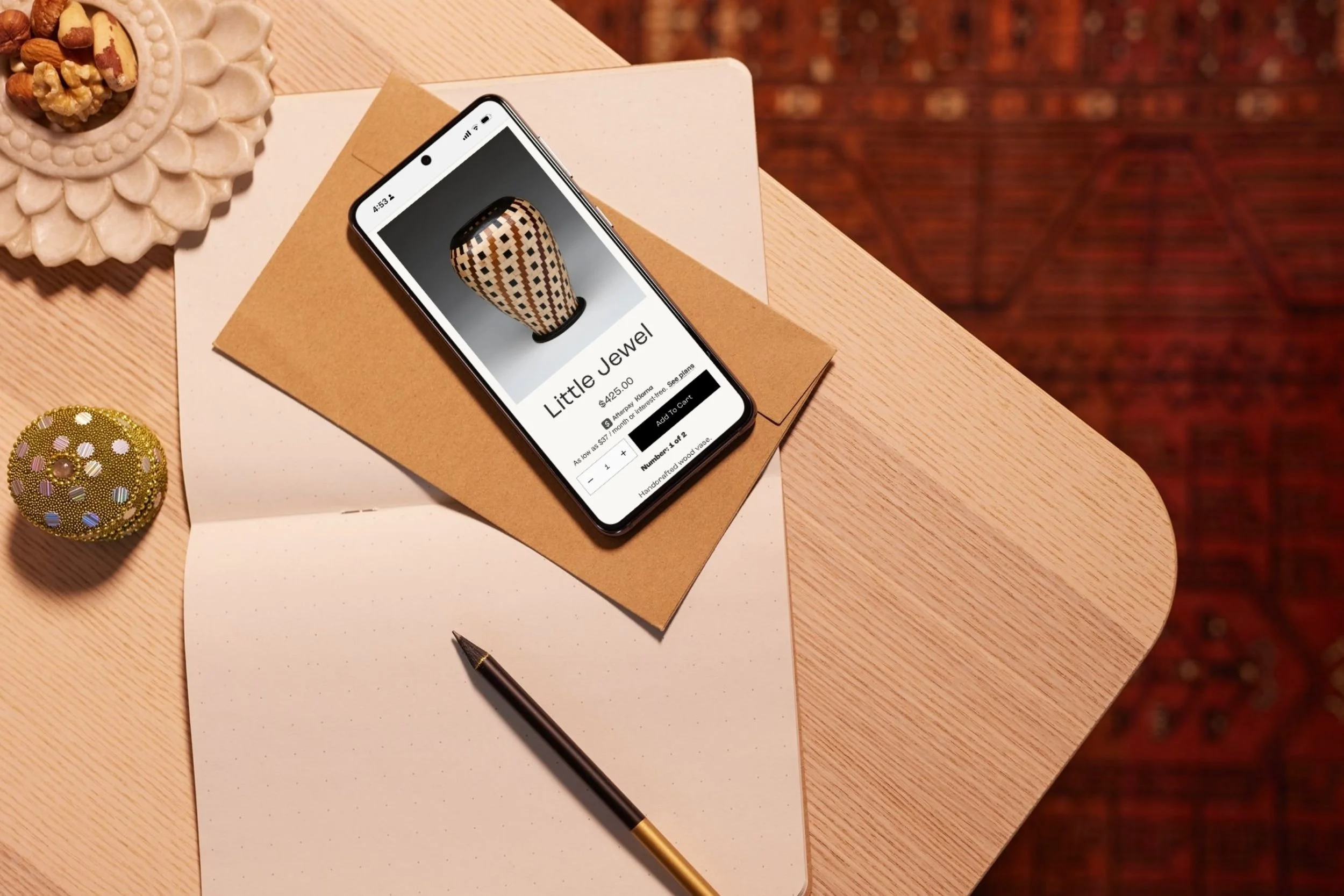 Smartphone on a wooden desk displaying an online jewelry store with a picture of a jewelry piece called Little Jewel priced at $425, surrounded by papers, a pen, a decorative bowl with nuts and dried fruit, a sequined ornament, and a plain white pape