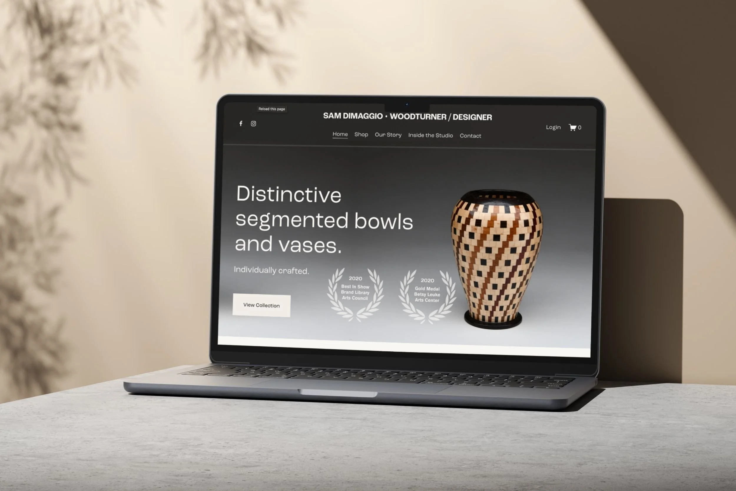 A laptop on a white table displaying a website about designer woodturner Sam DiMaggio. The website features a photo of a wooden segmented bowl or vase with a geometric pattern. Some blurred plant branches are visible on the left side of the image.