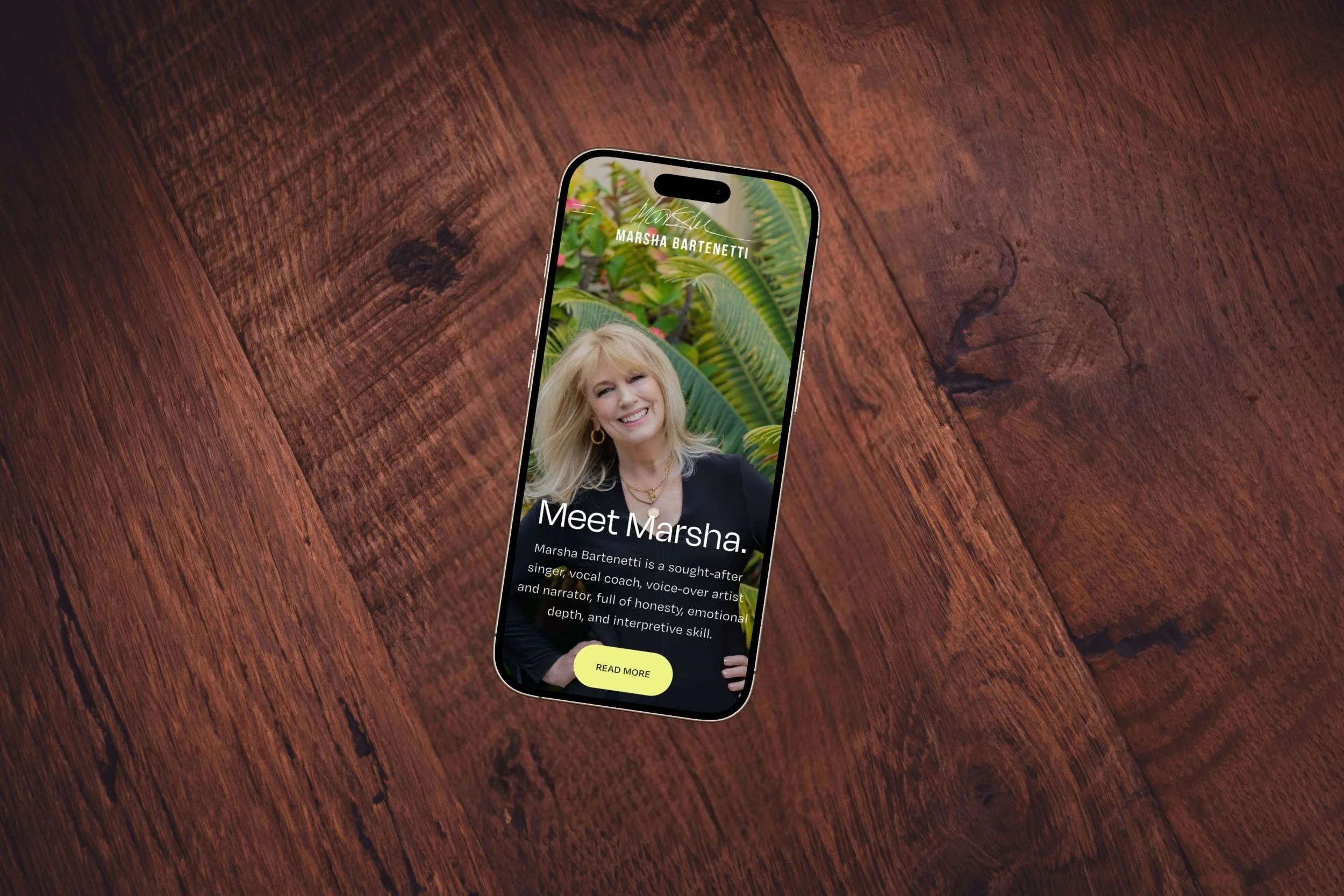 Smartphone on a wooden surface displaying a profile page for Marsha Bartenetti, with a photo of her smiling and green foliage in the background.