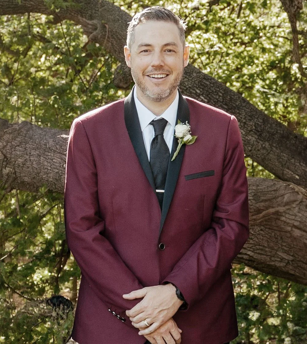 A man wearing a maroon tuxedo with black lapel, white shirt, black tie, and a boutonnière, standing outdoors in front of a tree with green leaves, smiling.