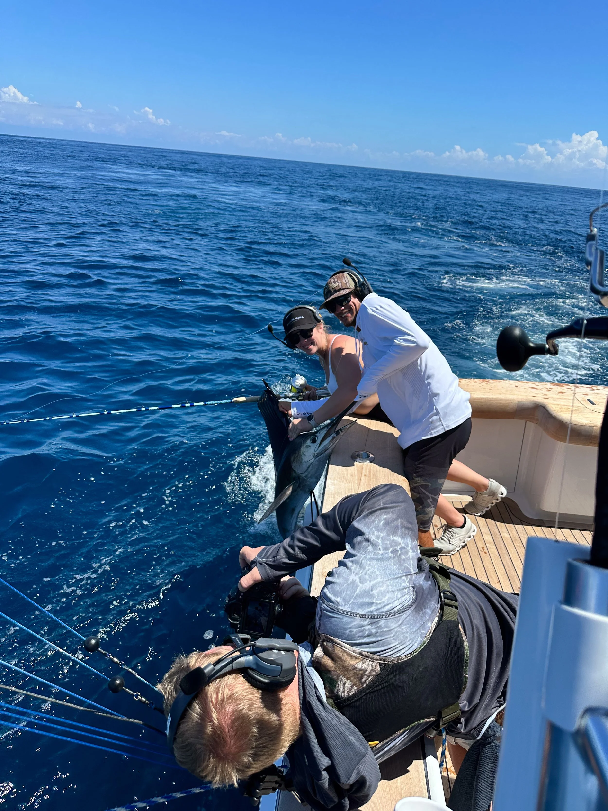 Two people on a boat holding a large fish, with the ocean and blue sky in the background, using fishing gear and wearing hats and sunglasses.