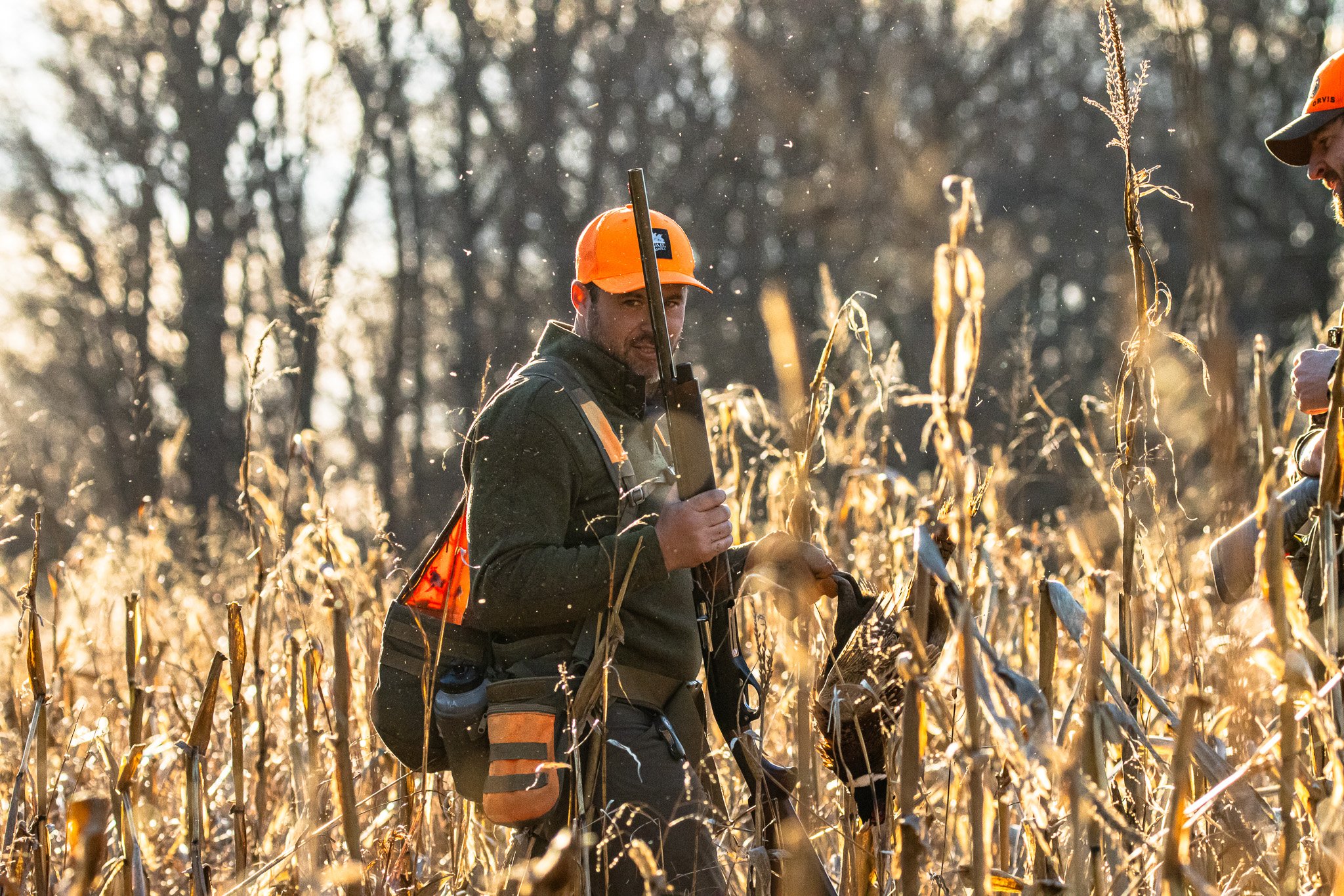 Two hunters, one man, and one woman, stand in a field of dried corn stalks during sunset, wearing orange safety gear and holding rifles.