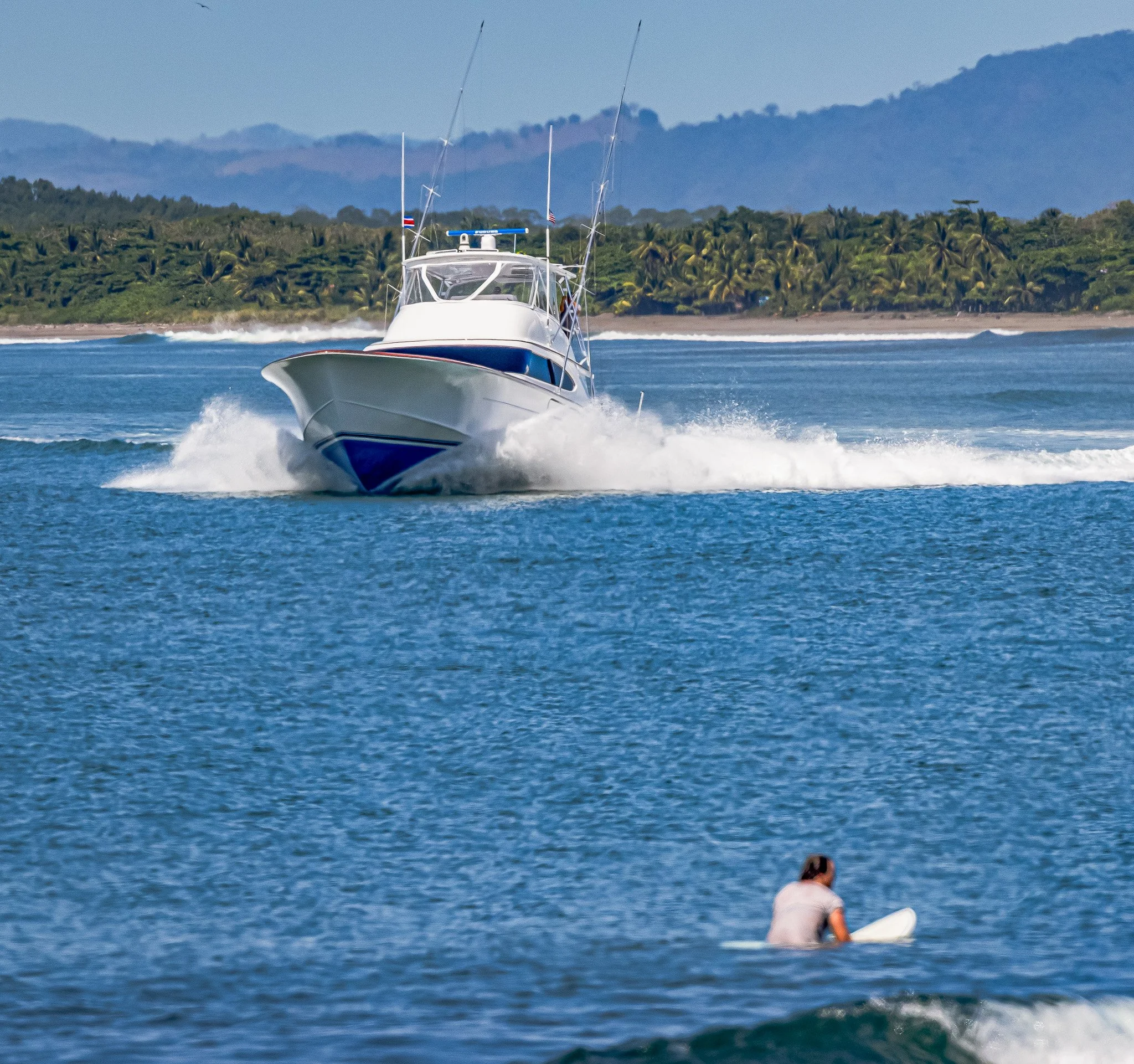 A white and blue motorboat speeding on the water near a tropical shoreline with lush greenery and mountains in the background, and a person sitting on a surfboard in the water.