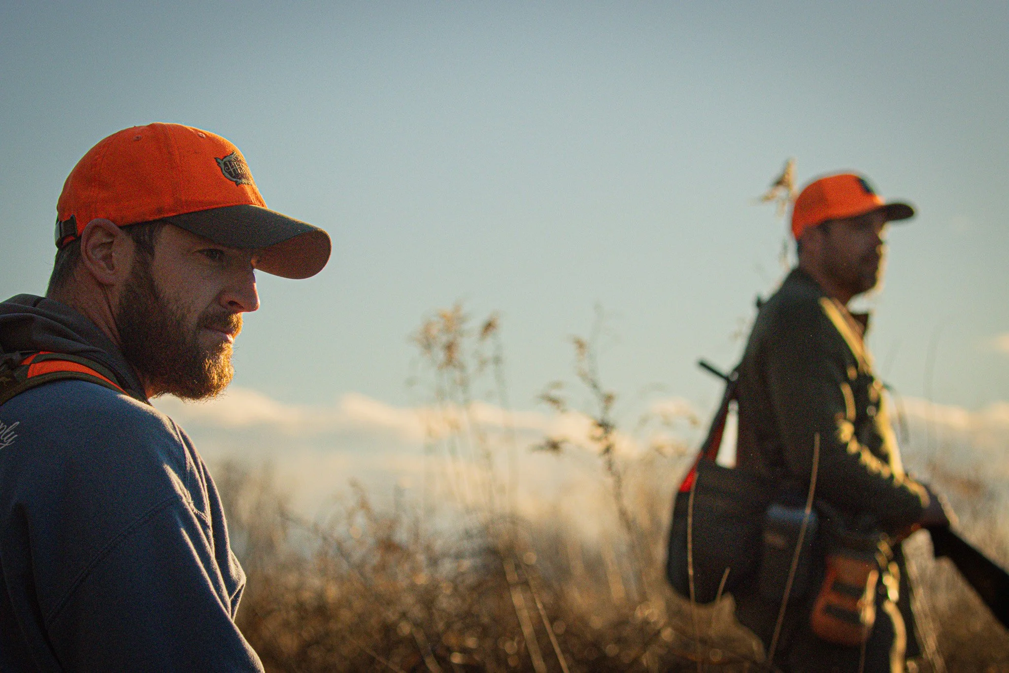 Two men wearing orange caps and dark clothing, standing outdoors at sunset or sunrise, with a blurred natural landscape and tall grasses in the background.