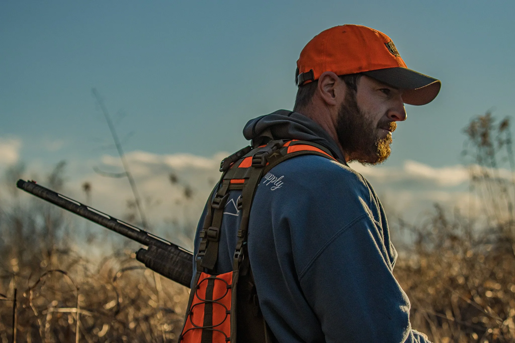 A man with a beard wearing an orange cap and a backpack, standing outdoors during sunset, with tall grass and a blurred background.