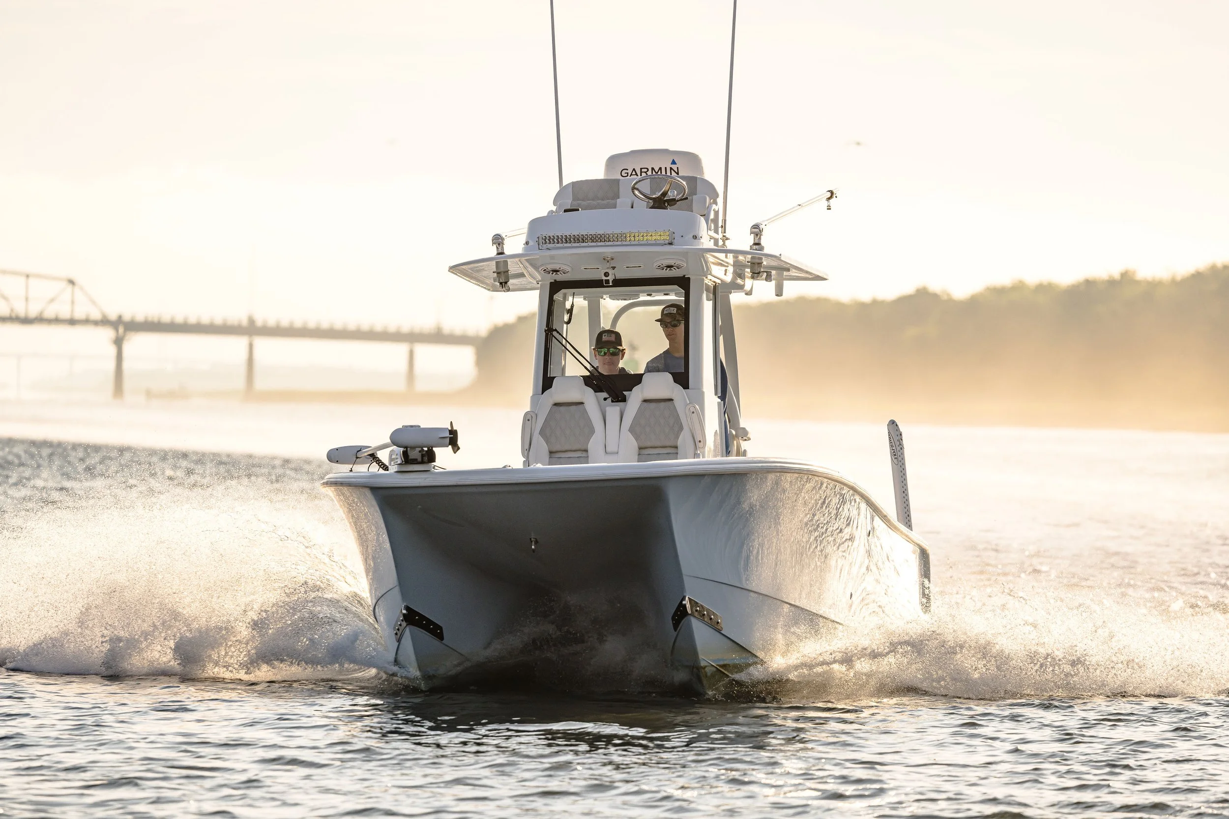 A white motorboat moving through water during sunset with two people onboard wearing sunglasses and caps. The boat has a Garmin radar and other equipment on top, and there is a bridge in the background.
