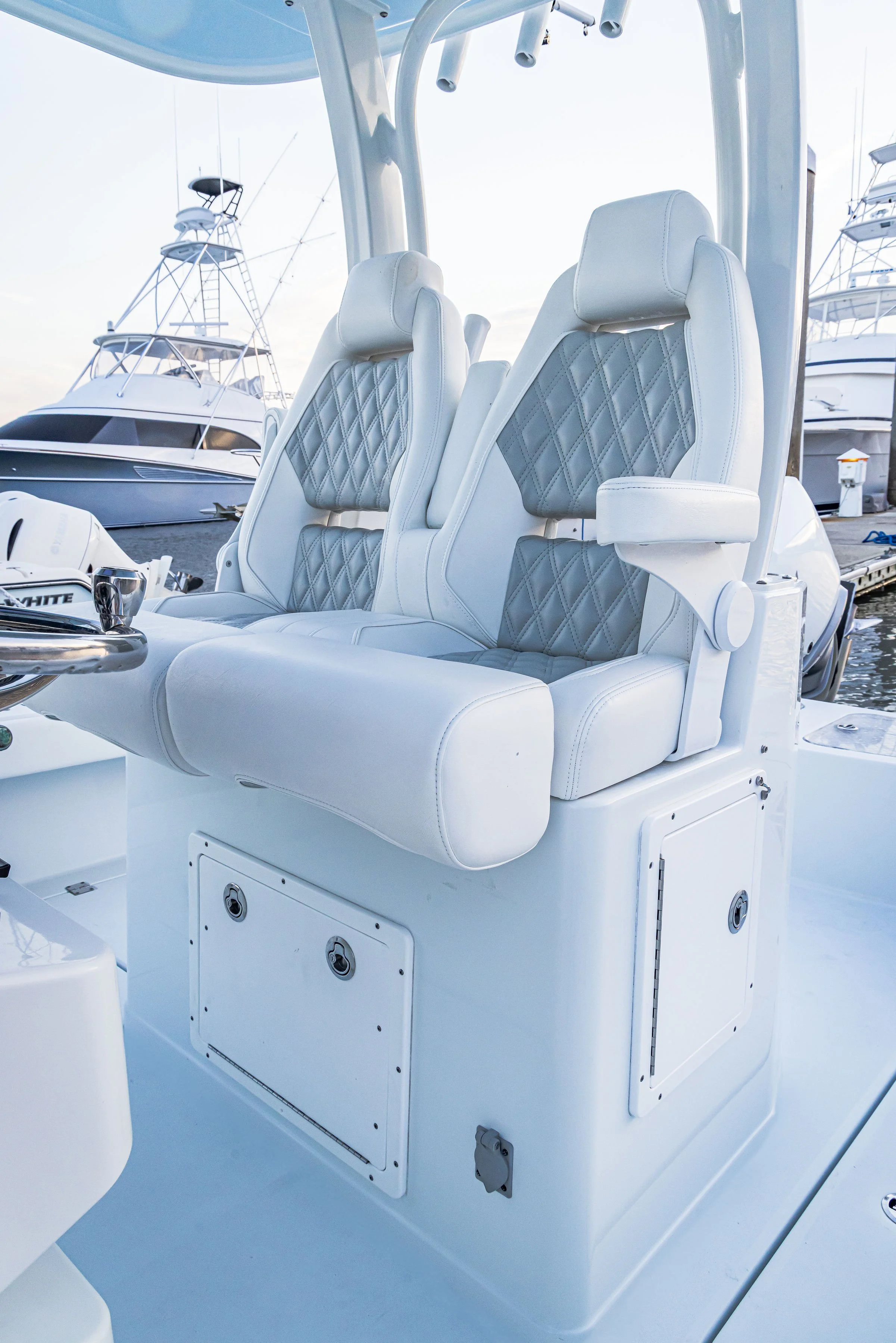 Close-up of a white boat's seating area with two cushioned seats, with a marina and yachts in the background.