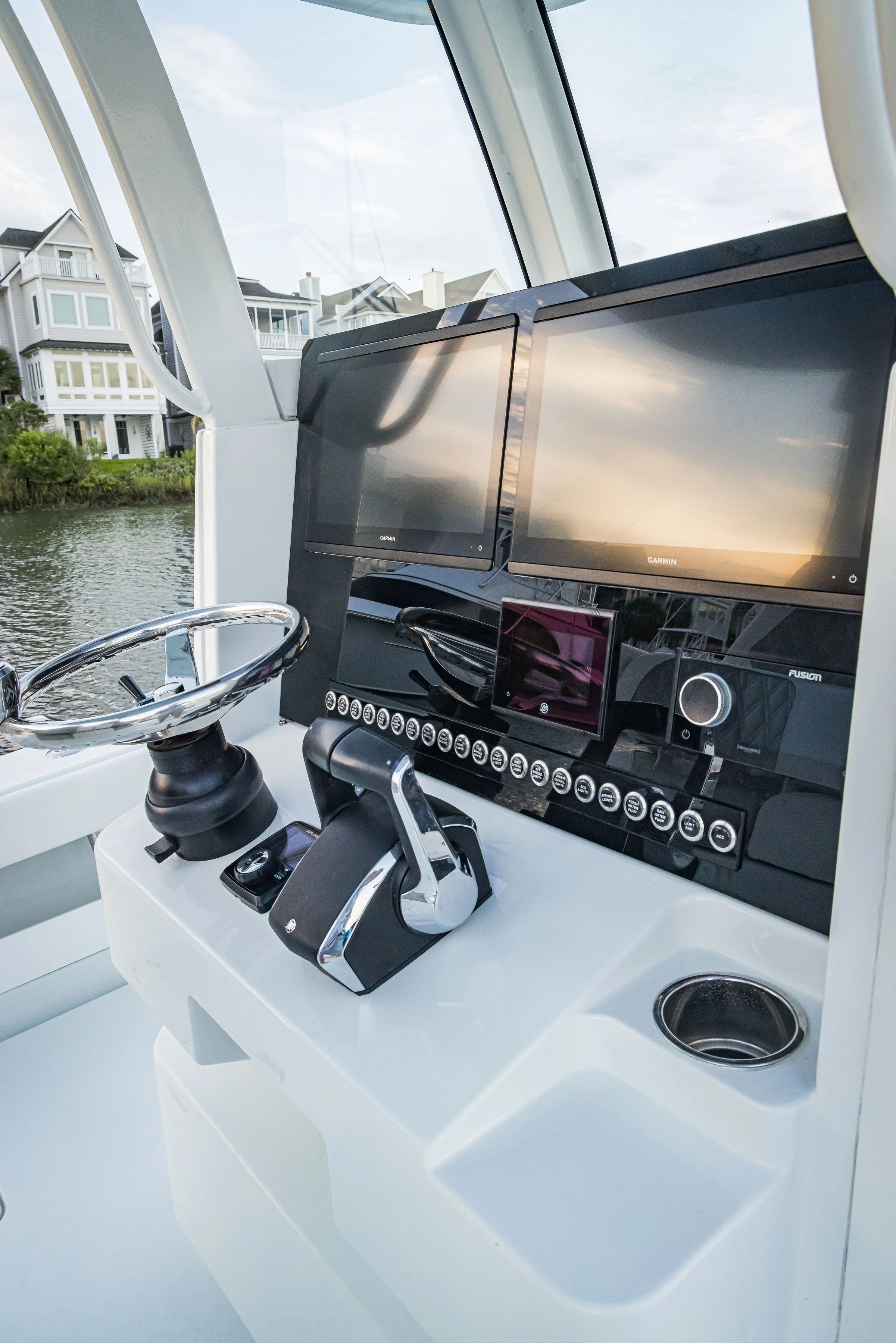 The image shows the helm of a boat with two Garmin screens, a steering wheel, and other controls. Behind, there are modern houses along a waterway, suggesting the boat is near a residential area.