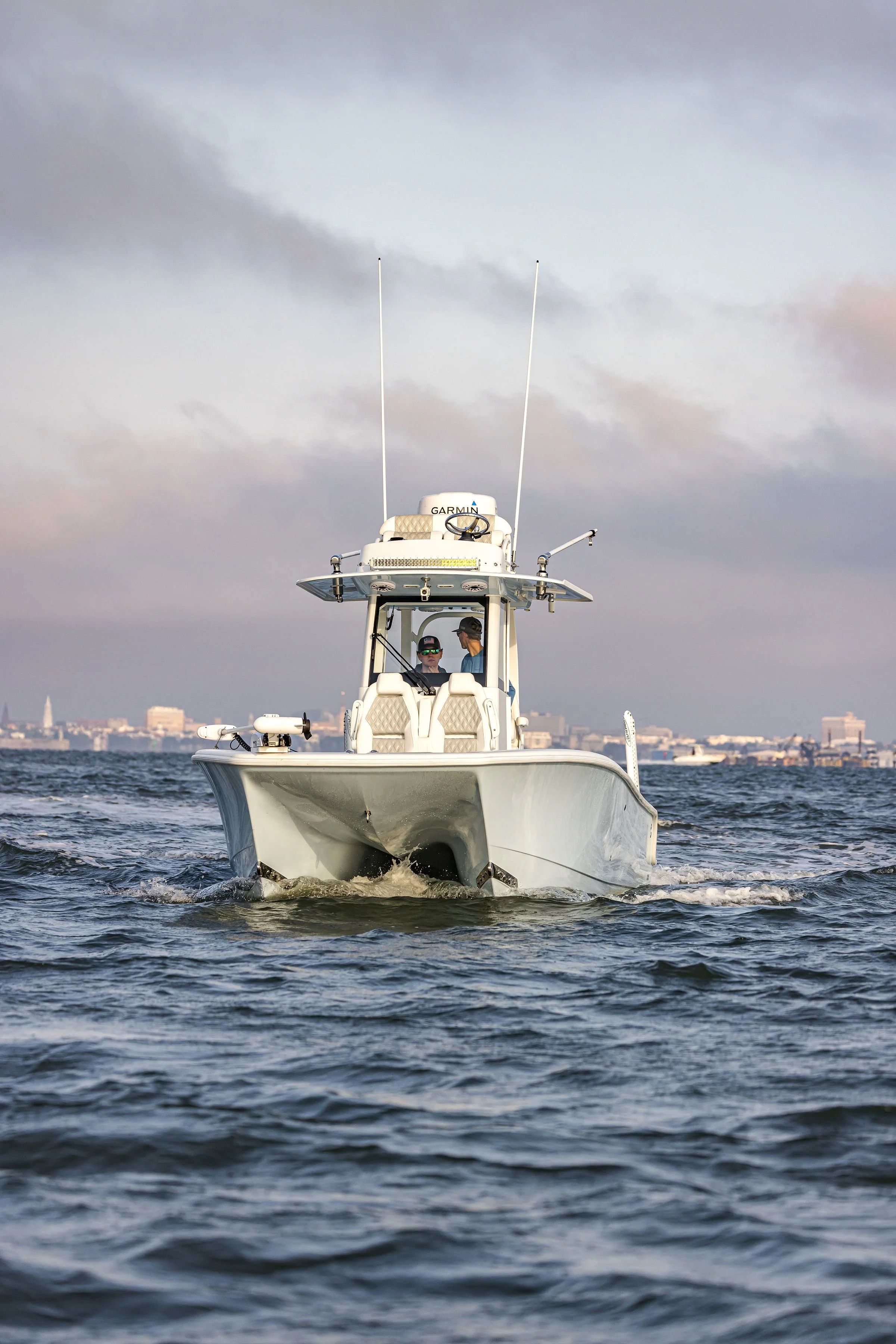 A white boat is on the water with the city skyline in the background, and a person wearing sunglasses and a hat is on the boat.
