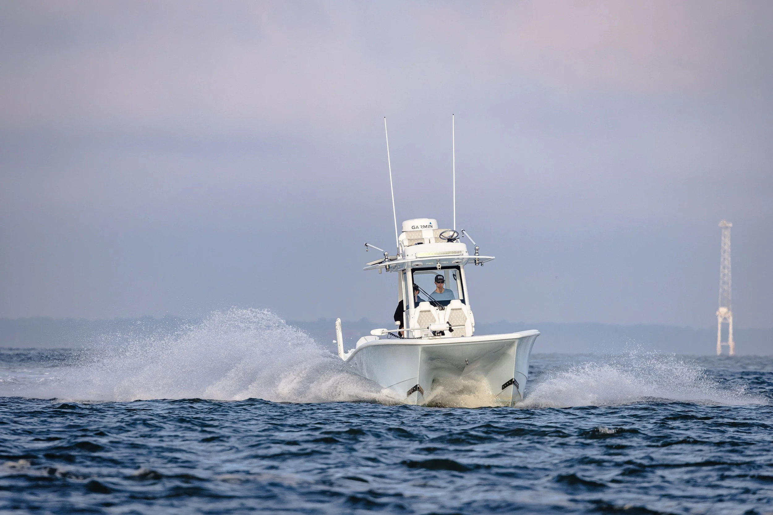 A person driving a white motorboat on the water, with a structure resembling a communication tower in the background.