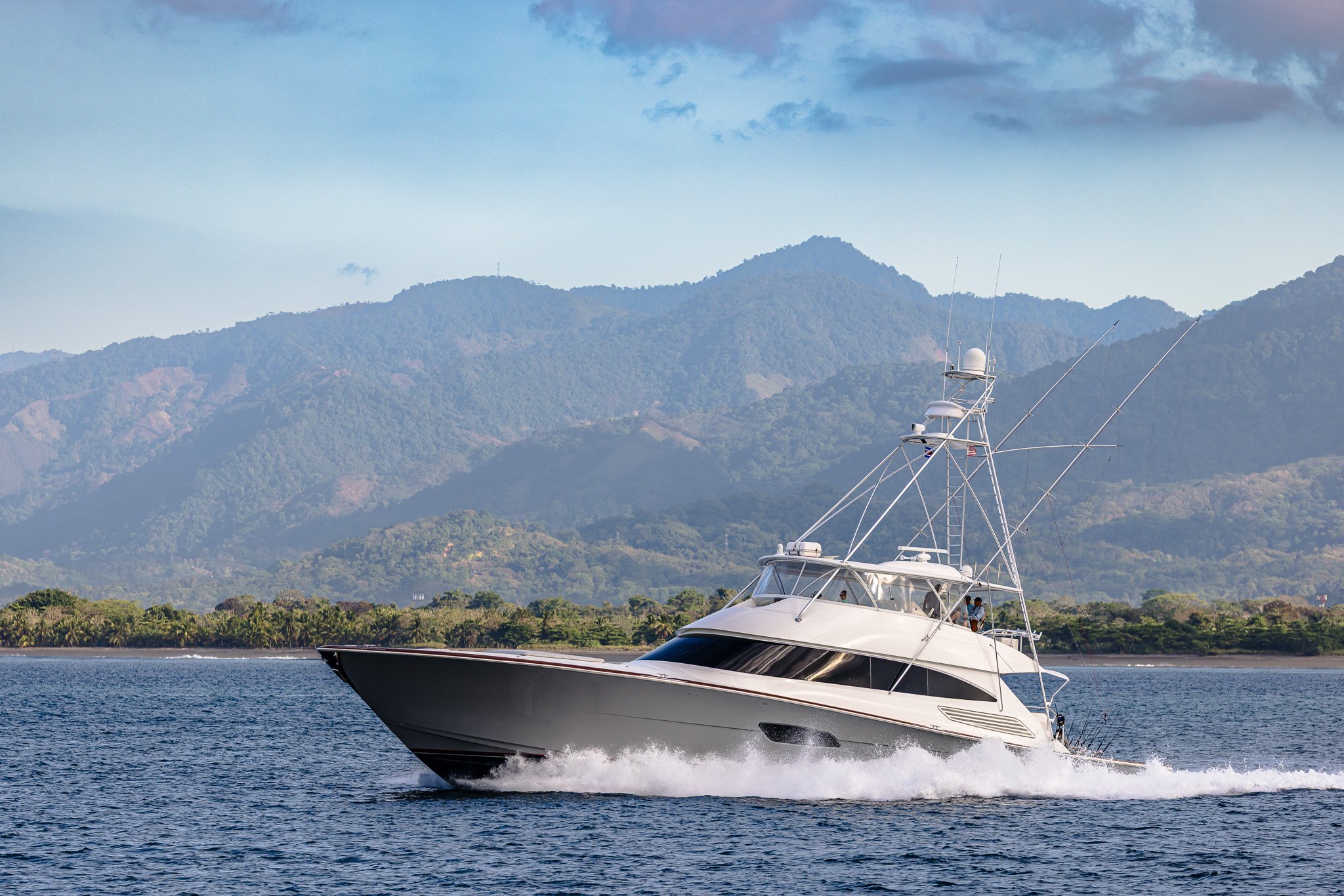 A white yacht cruising on the water with a mountain range in the background and a partly cloudy sky.
