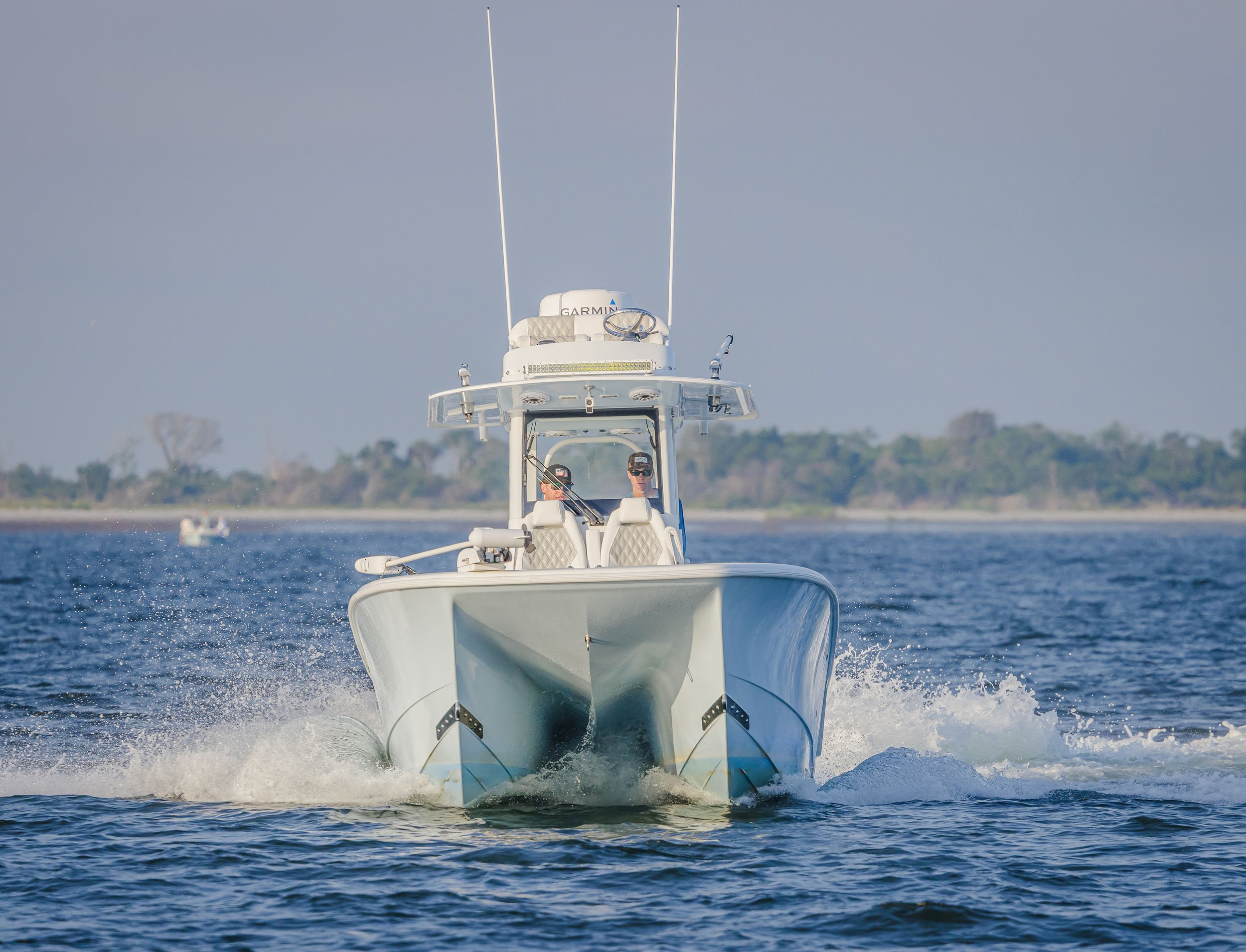 A white motorboat moving fast on the water with two people on board, and a faint shoreline with trees in the background.