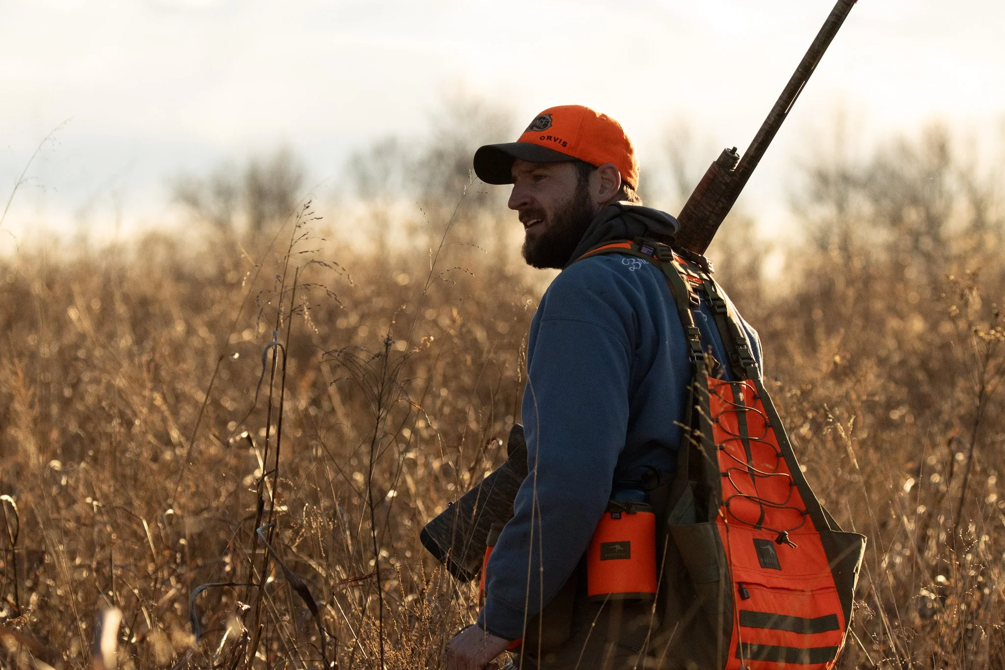 A man wearing an orange cap and blue jacket stands in a field of tall, dry grass during sunset, carrying a rifle over his shoulder and a backpack with orange accents.