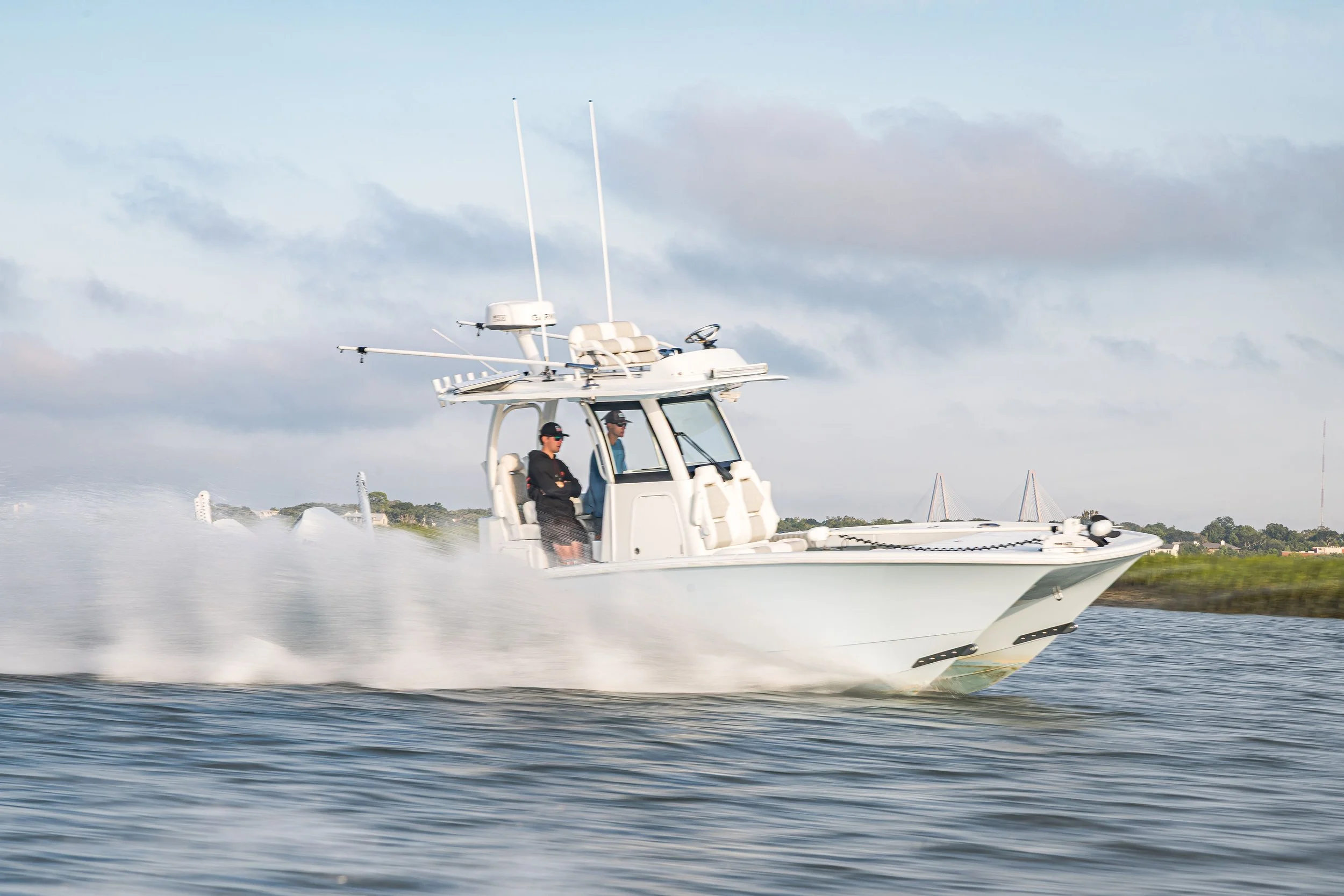 A white fishing boat moving quickly on the water with two men onboard, one standing and the other sitting, under a partly cloudy sky.