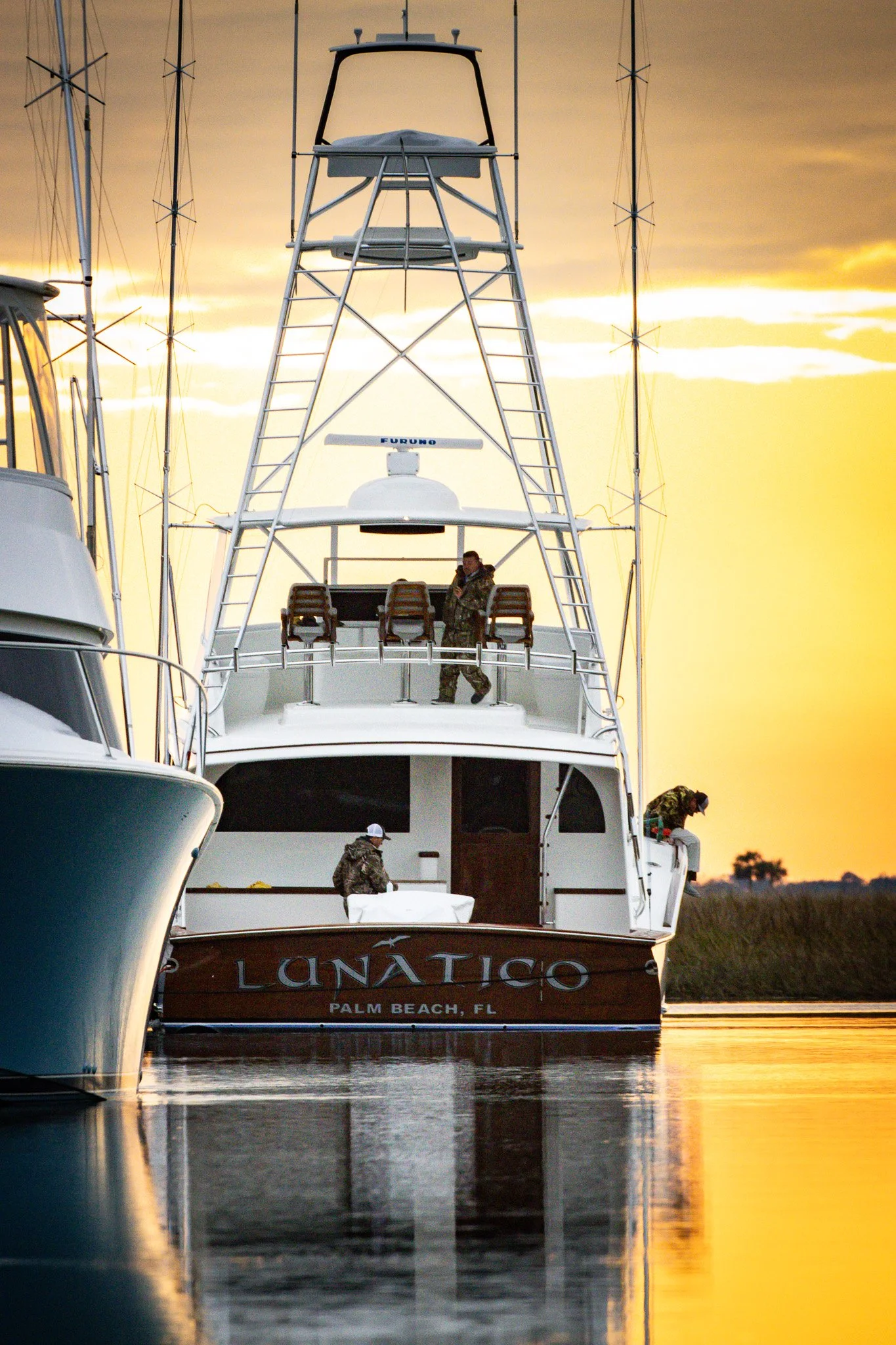 A large yacht with the name 'Luna Tico' and home port 'Palm Beach, FL' docked at a marina during sunset. Two people are on the upper deck, one standing and one sitting, with the background showing a golden sky and calm water reflecting the yacht.