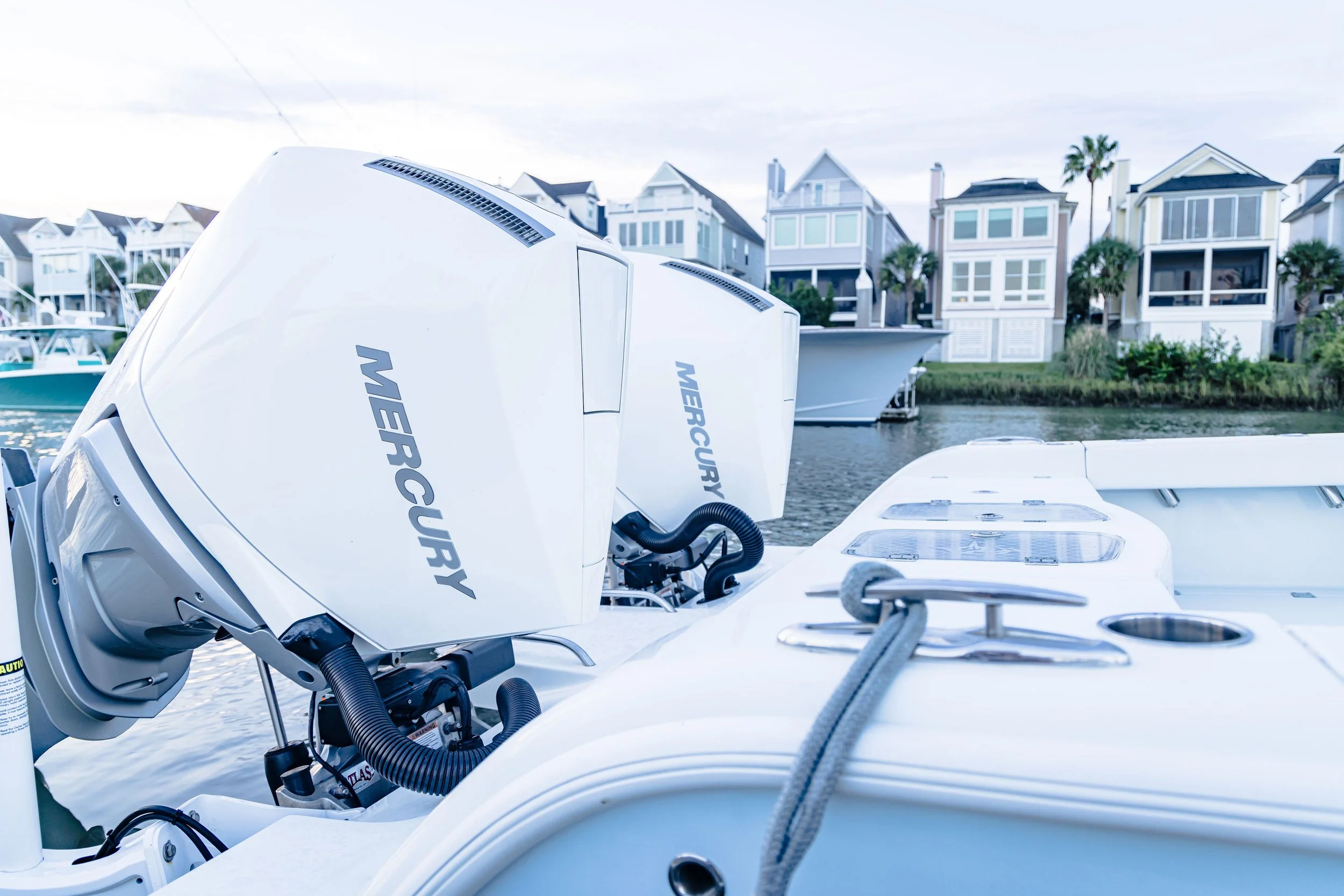Close-up of Mercury outboard motors on a boat, with a row of colorful houses and palm trees in the background.