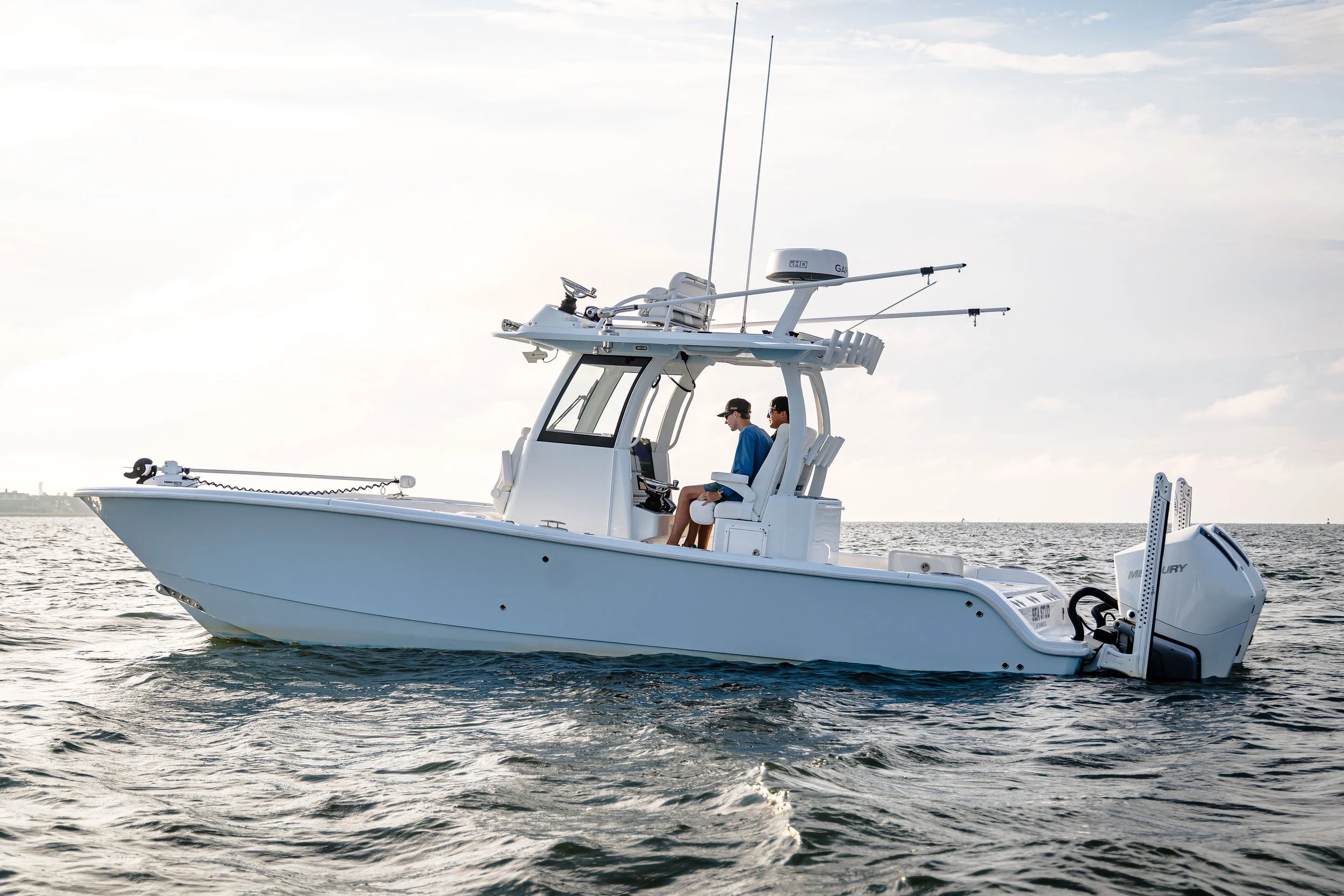 A white fishing boat with two people sitting inside, floating on water under a partly cloudy sky, equipped with nautical gear and electronics.