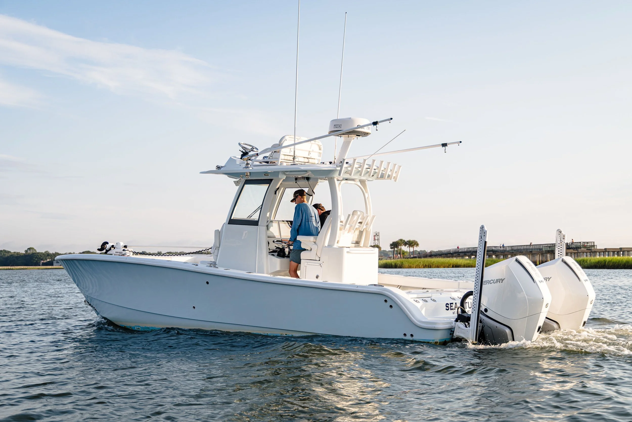 A white fishing boat on the water with two men aboard. One man stands near the helm, and the other sits inside the boat. The boat has multiple antennas and fishing rod holders on the roof. In the background, there is a shoreline with trees, a bridge,