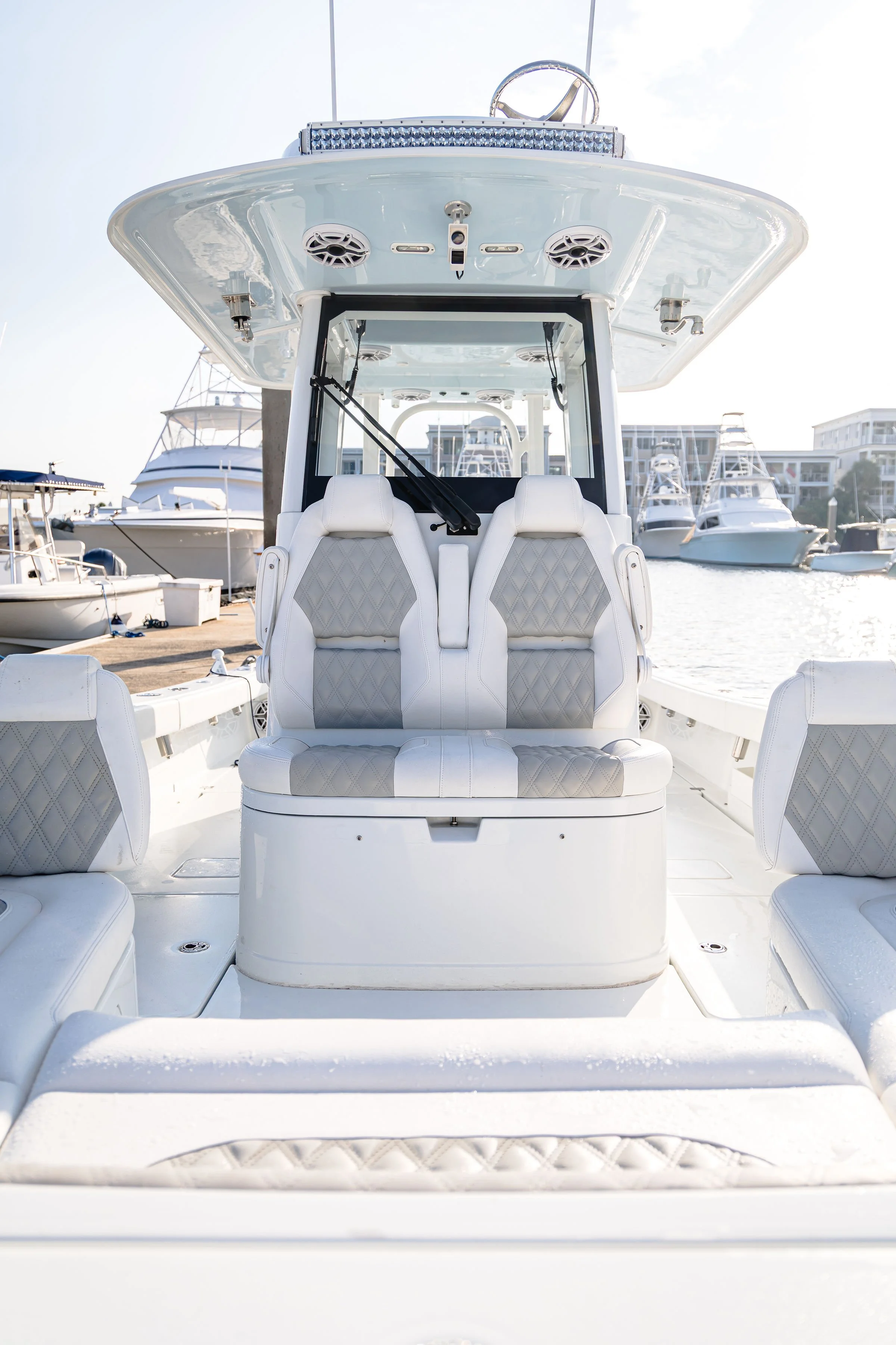 View of a white yacht's cockpit with cushioned seats, steering wheel, and boat hardware, docked at a marina with other boats and buildings in the background.