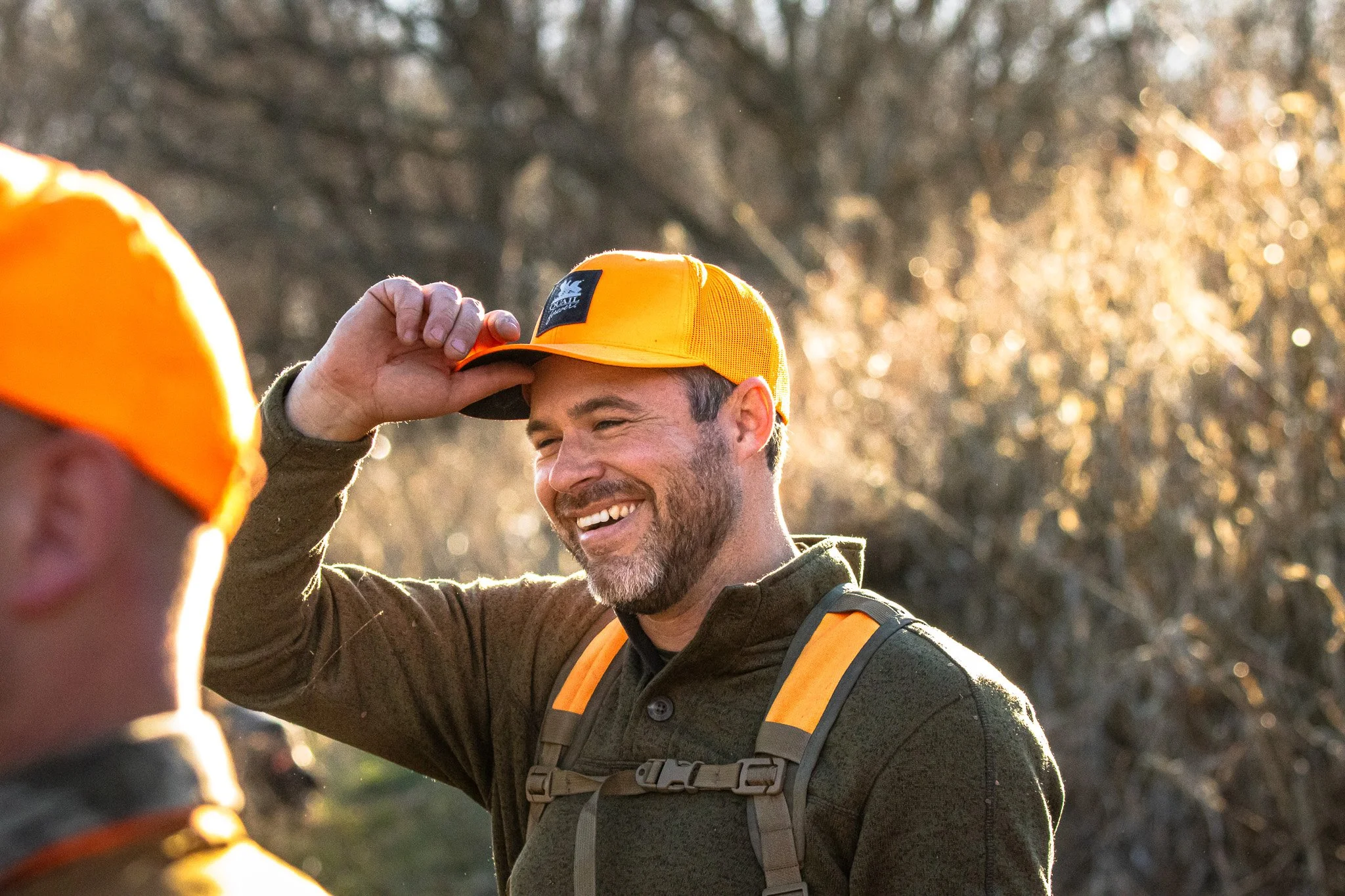 A man smiling and wearing a yellow cap and outdoor gear, adjusting his cap. The background shows trees with sunlight filtering through.