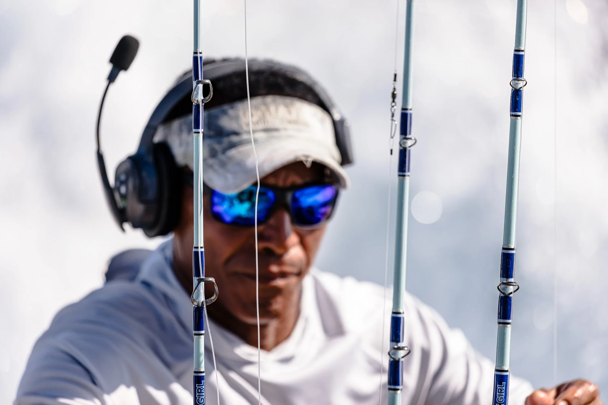 A person wearing sunglasses, a cap, and a white jacket, standing outdoors during the daytime, is surrounded by fishing rods with blue details.