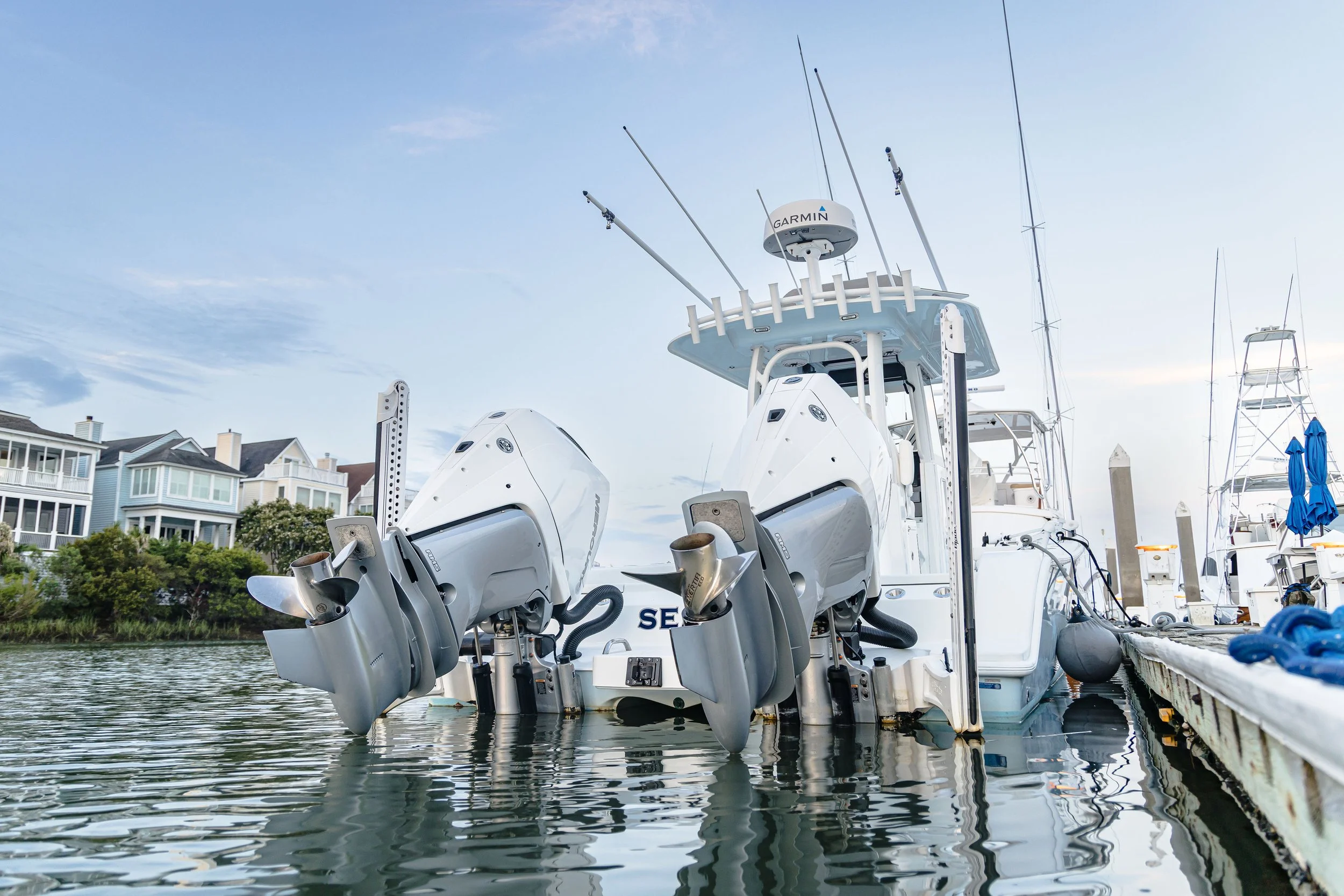 View of yachts docked at a marina with multi-story coastal houses in the background, under a partly cloudy sky.