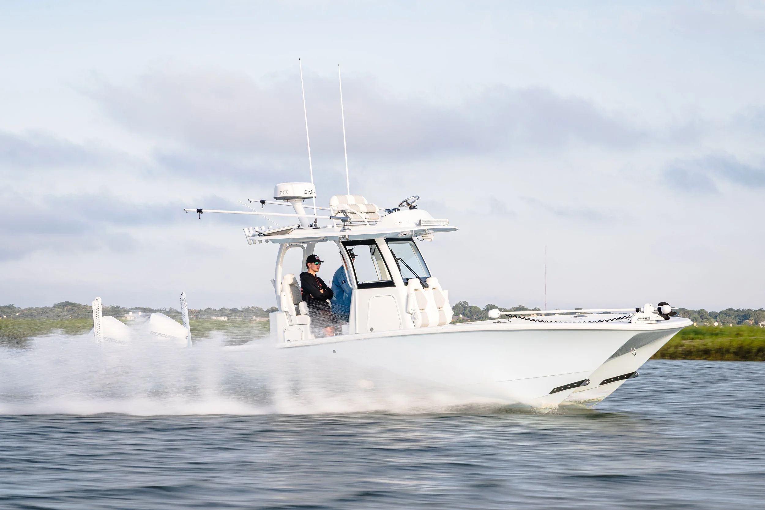 A person operating a white motorboat moving quickly on the water, with a cloudy sky and distant shoreline in the background.