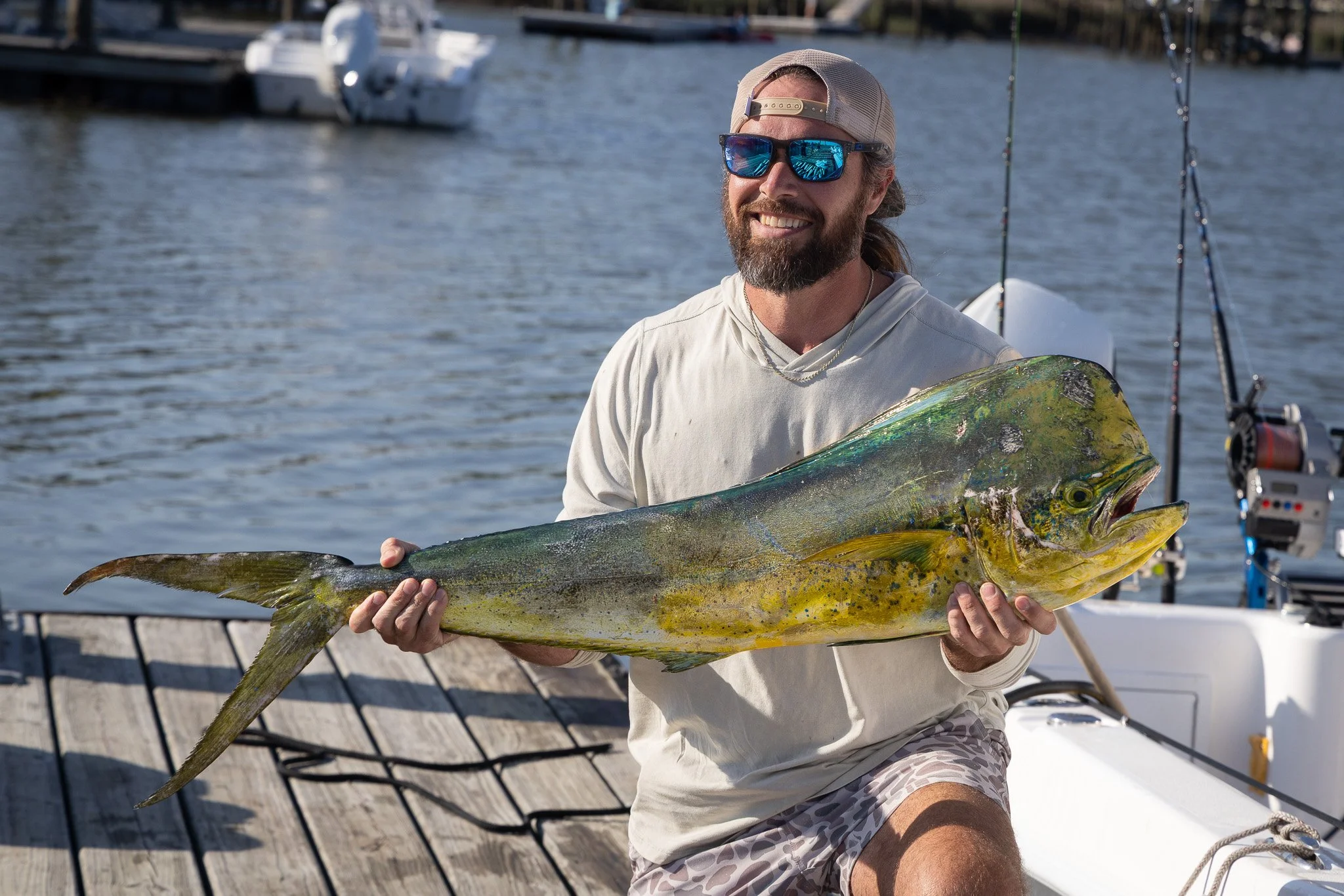 A man with long hair, beard, sunglasses, and a baseball cap is sitting on a boat holding a large fish, likely a yellowtail or amberjack, with a marina and boats in the background.