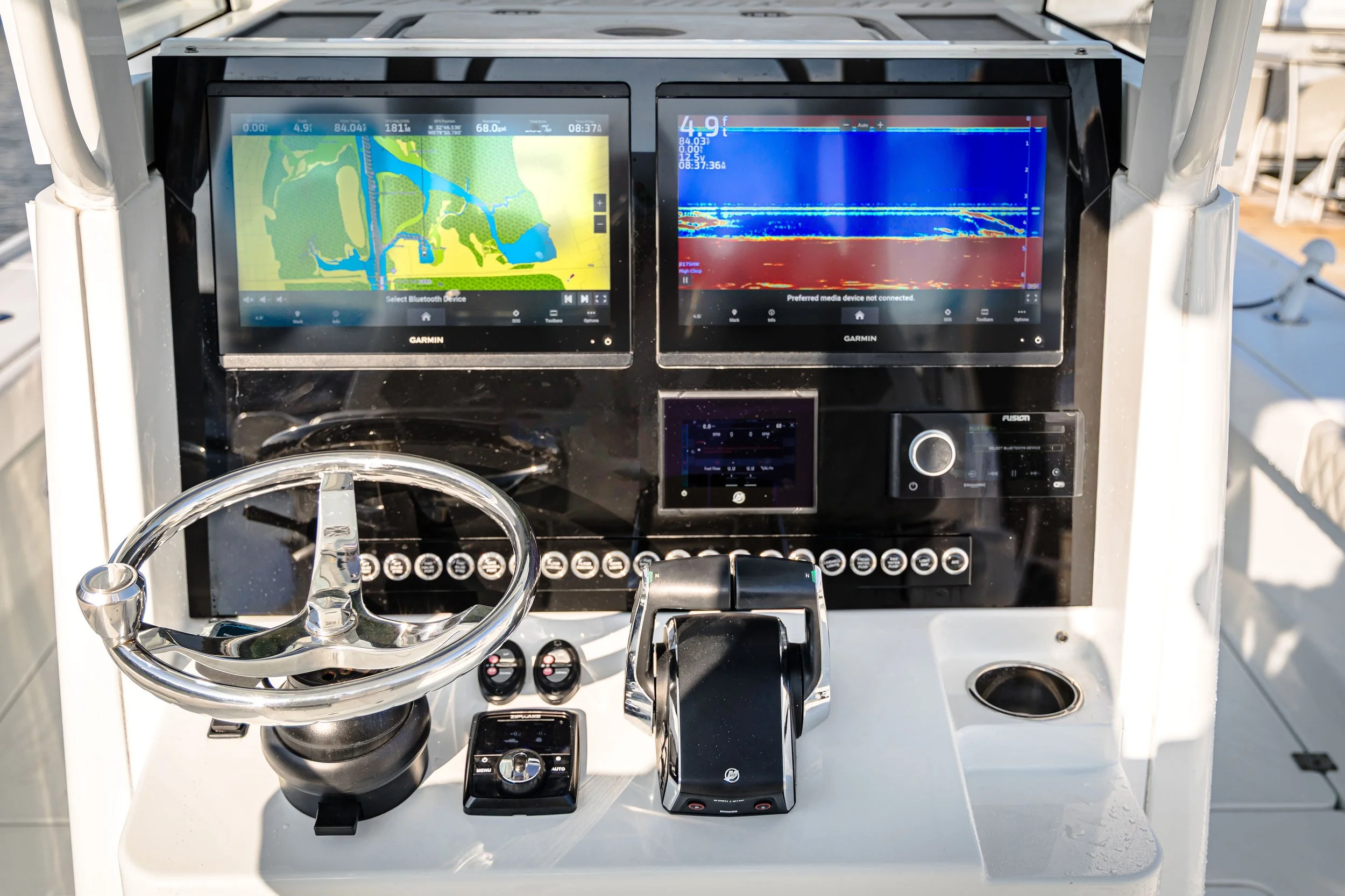 Close-up of a boat's control panel with two Garmin screens displaying navigation and sonar data, a steering wheel, and various switches and electronic devices.