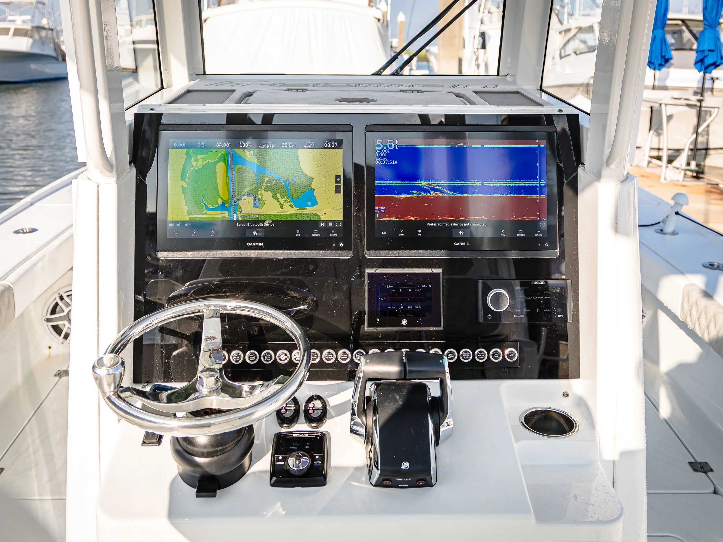 Close-up view of boat control console with two Garmin screens displaying navigational and sonar data, along with steering wheel, throttle, and other controls, on a marina dock.
