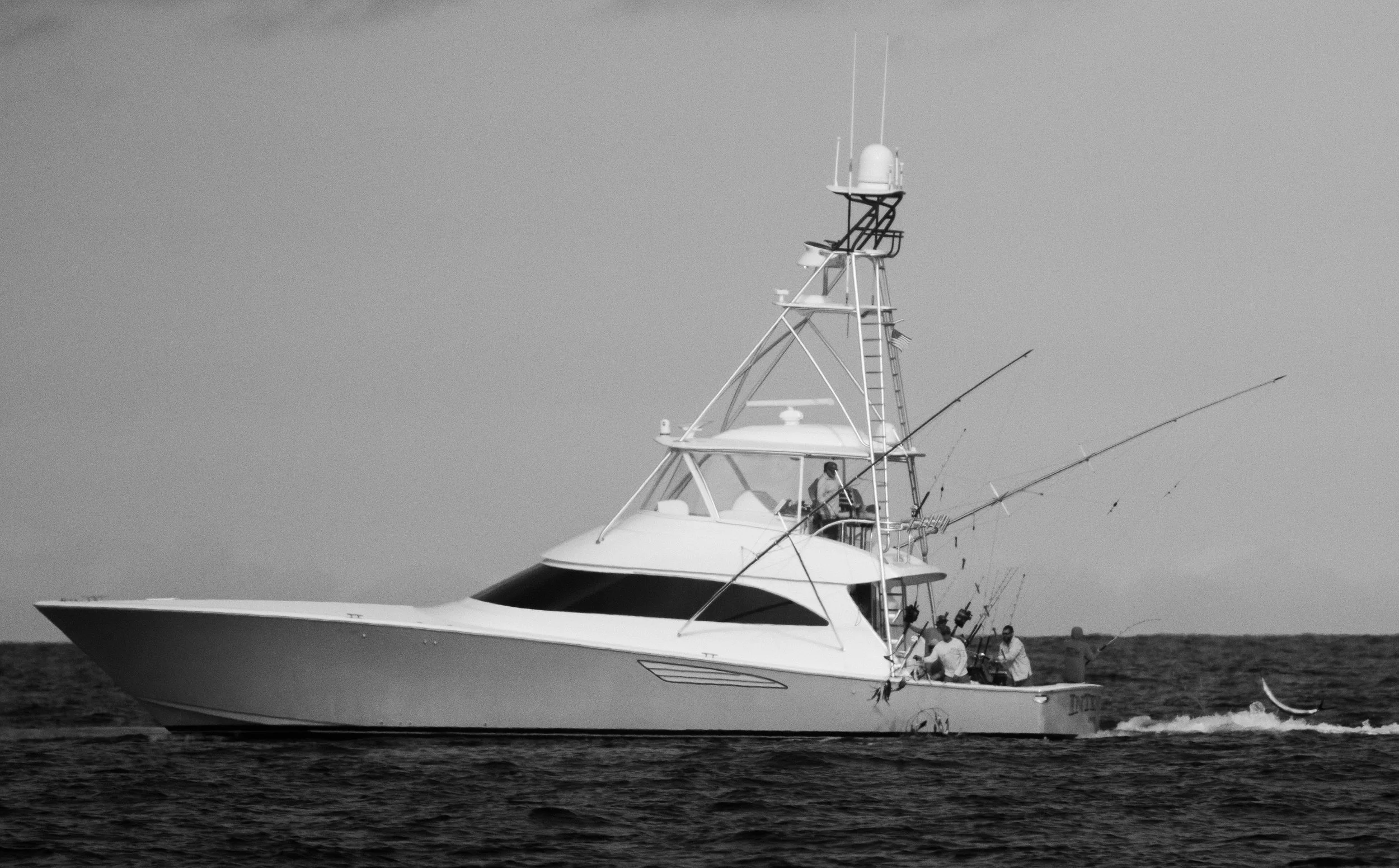 A black and white photo of a large yacht with several people on deck, fishing rods, and communication antennas on top, on the water with a distant shoreline.