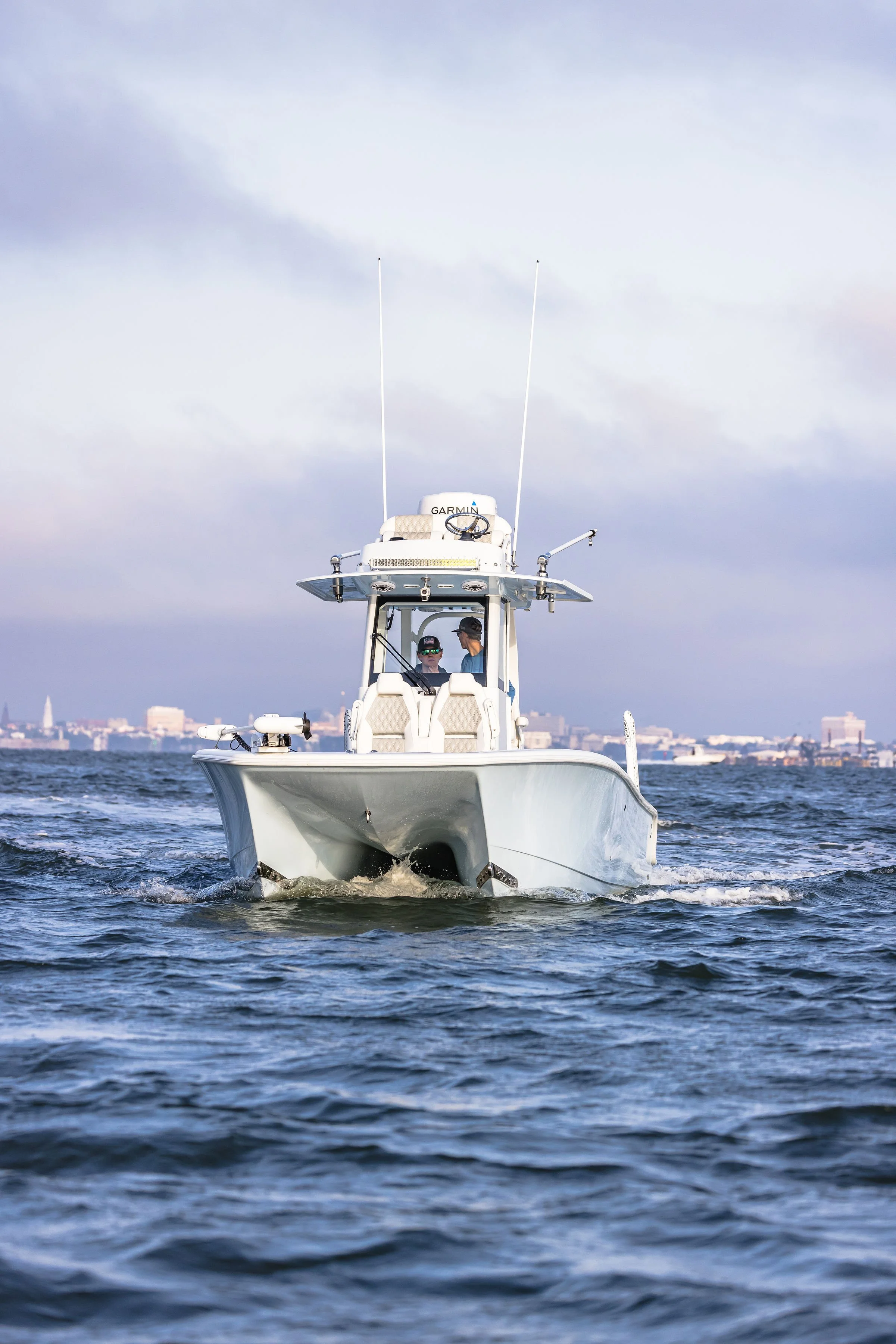 A white boat with two people on it sailing on the water near a city, with a city skyline in the background under a partly cloudy sky.