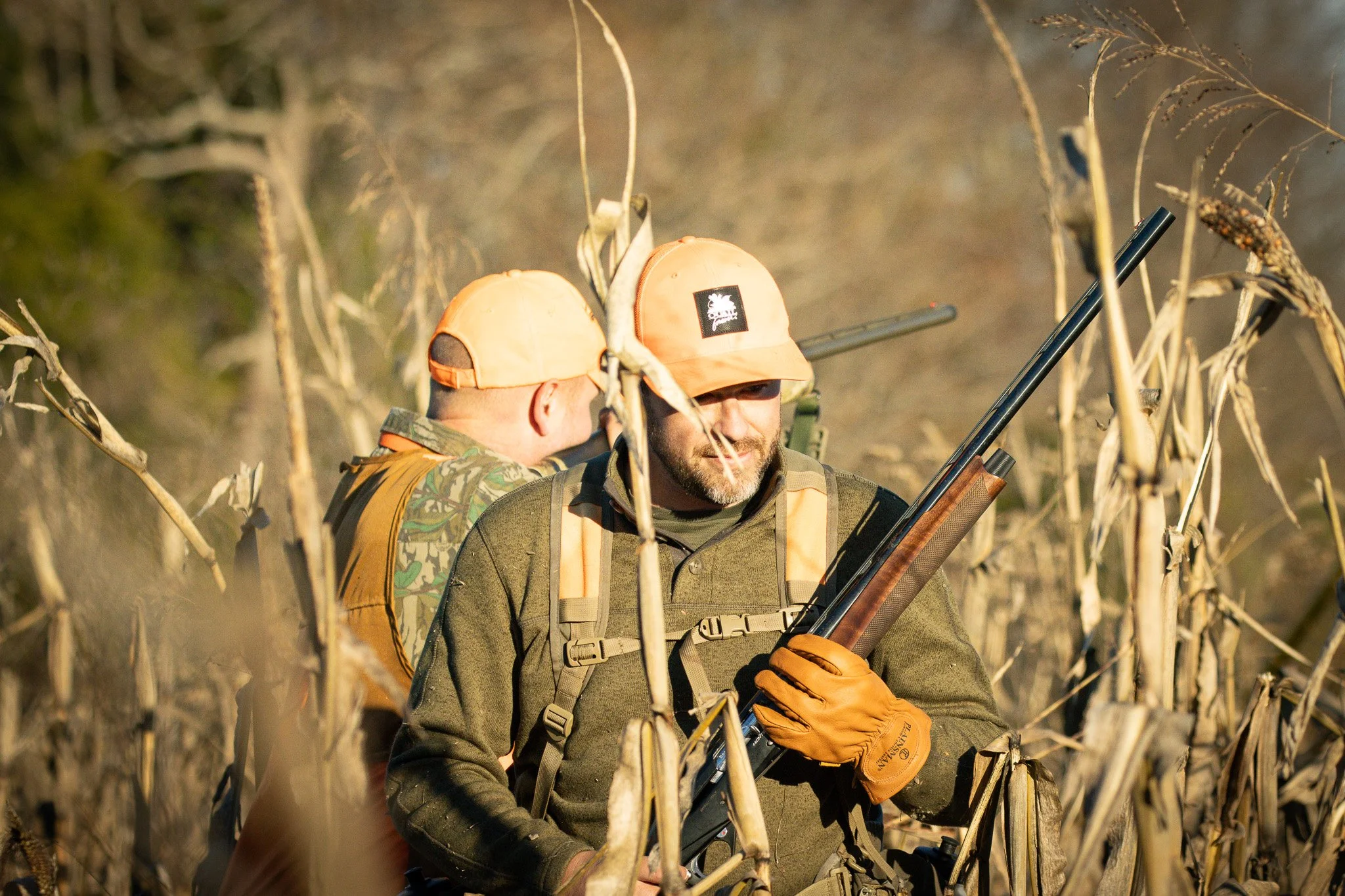 Two men in camouflage and orange hats are in a field of dried plants with a rifle. One is facing away, the other is smiling and looking to the side.