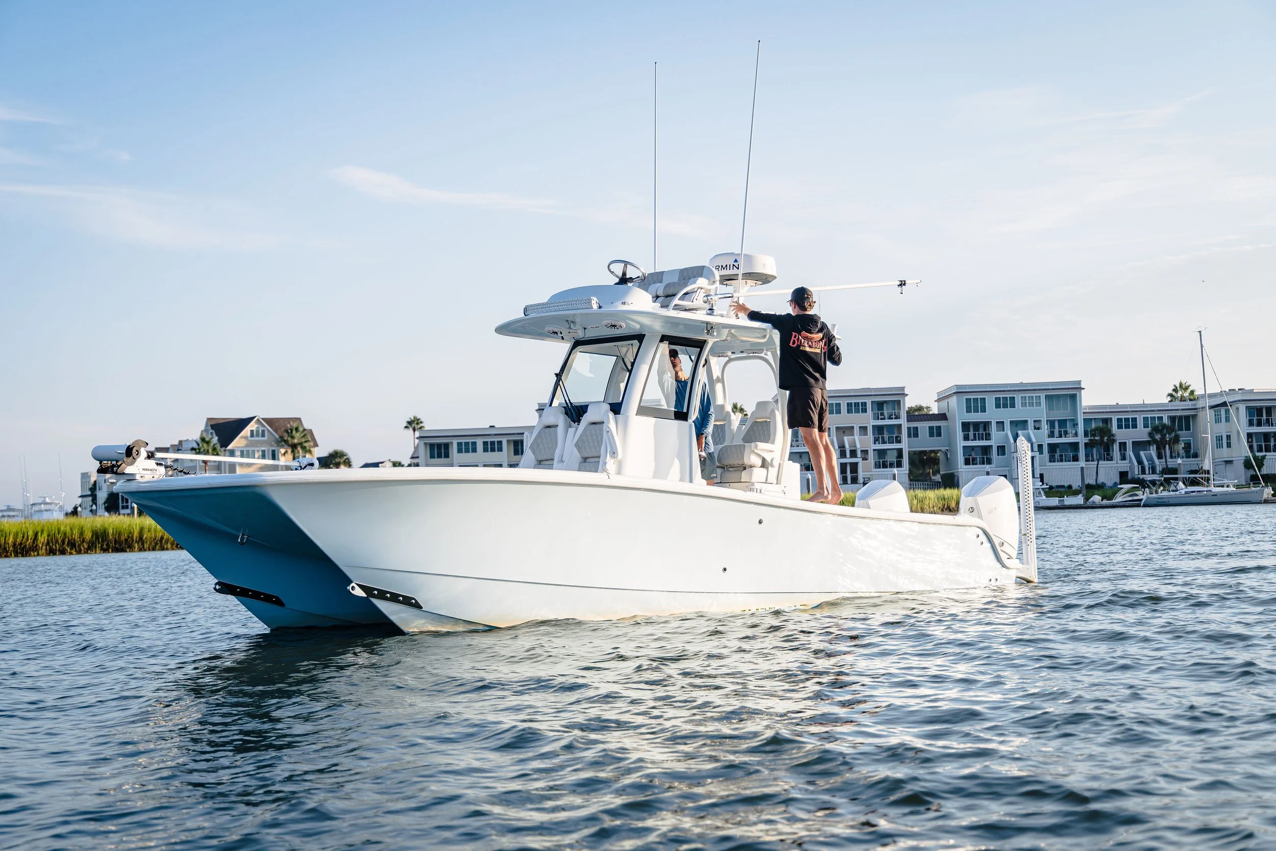 A person standing on a white motor yacht, adjusting equipment on the roof, with a marina and residential buildings in the background during daylight.