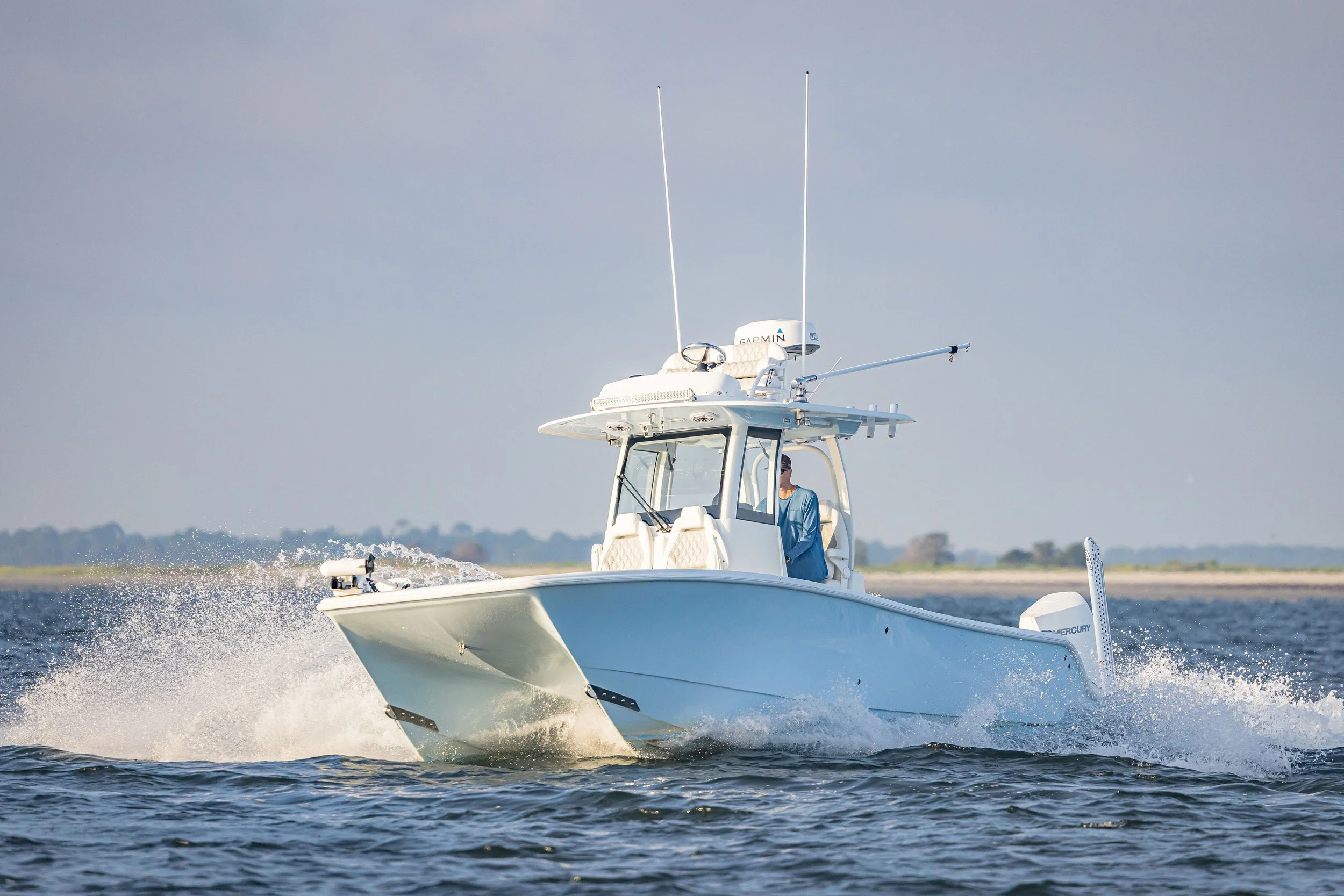 A white boat with a person in a blue shirt sailing on the water, equipped with radar and antennas, creating a wake as it moves through the water under a cloudy sky.