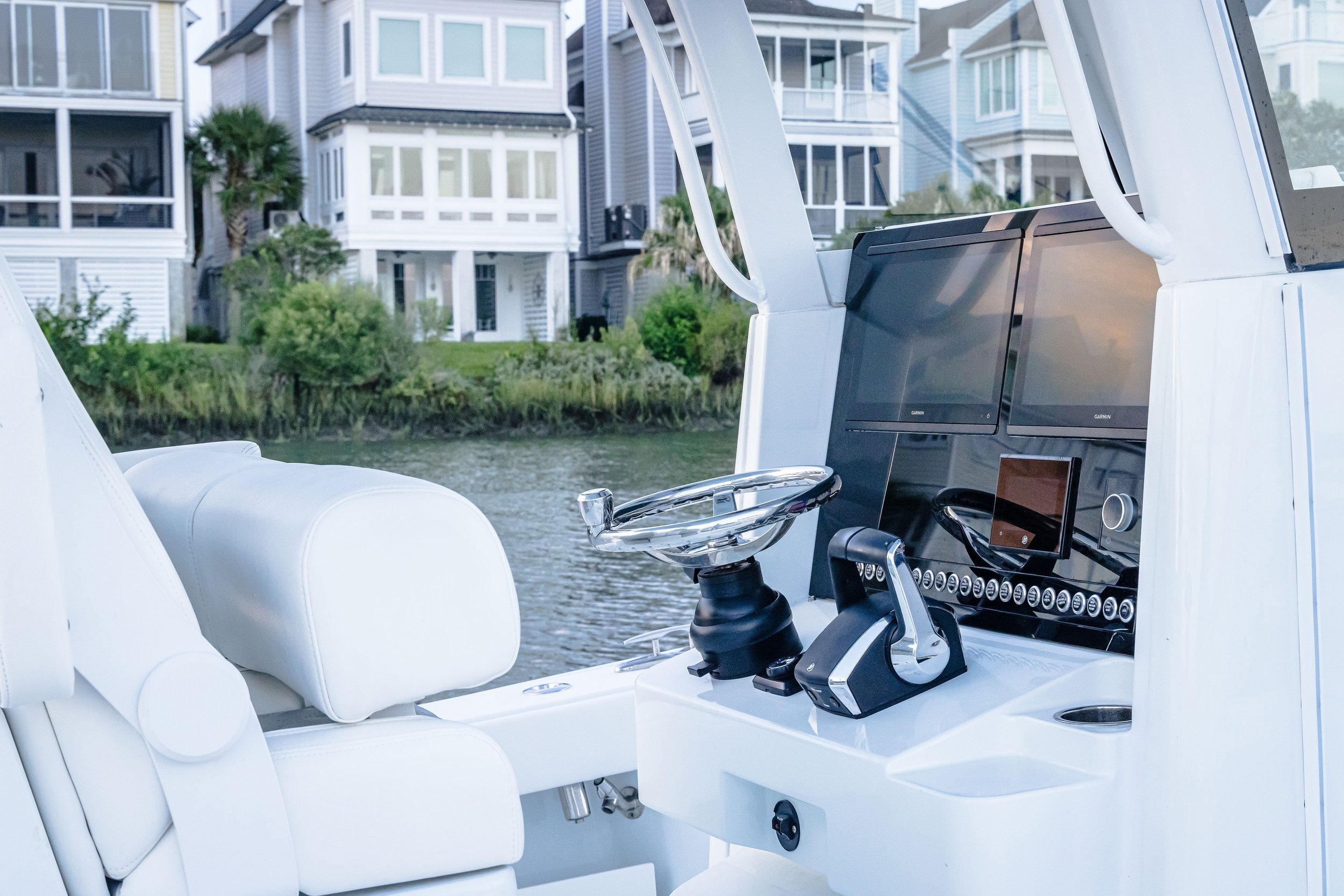 Close-up of the helm and dashboard of a white boat with a large flat-screen displaying navigation or sonar information, with houses and greenery in the background near the water.