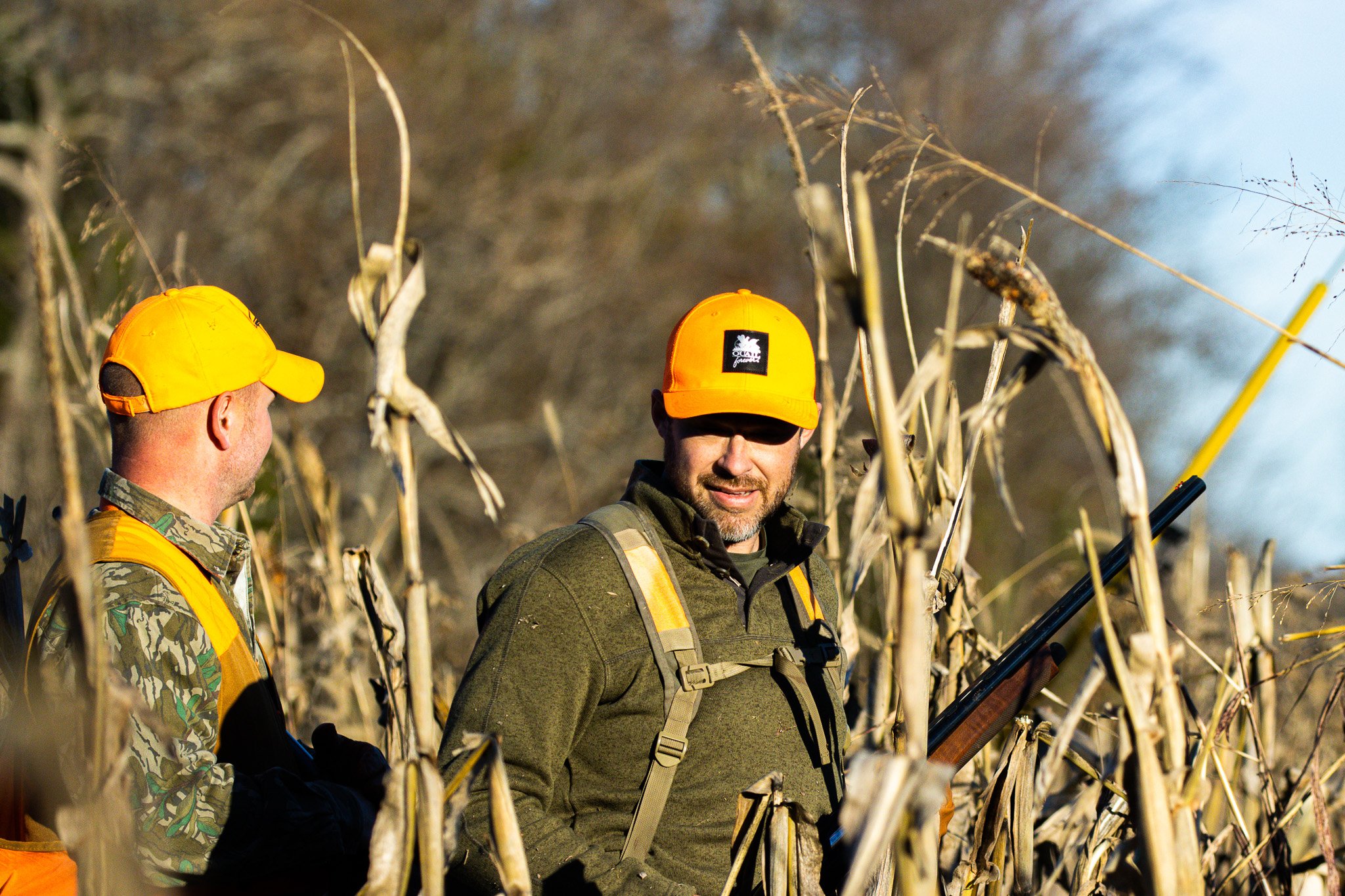 Two men wearing camouflage and bright yellow caps, standing among dry corn stalks in a field, with one of them holding a tool or rifle, having a conversation.