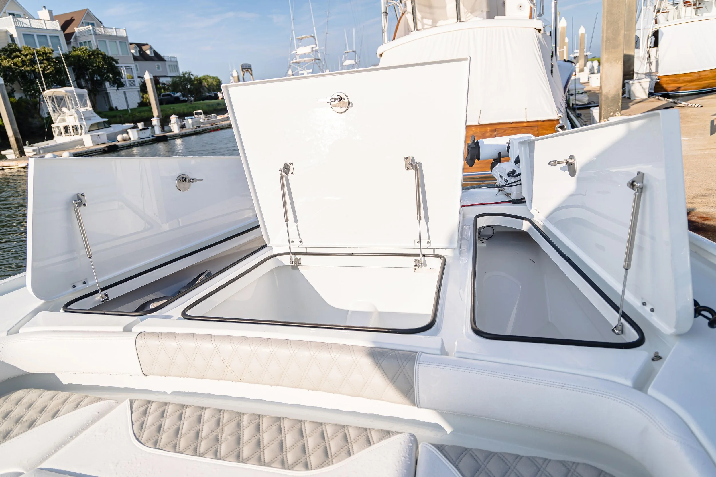 Close-up view of a white boat's interior with built-in seating, storage compartments, and hydraulic support rods, docked at a marina with other boats and residential buildings in the background.