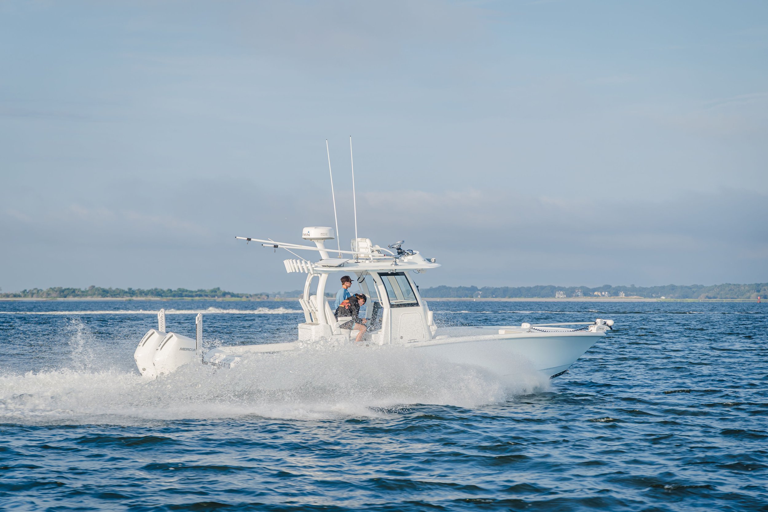 A white powerboat speeds across the water with two people onboard, one steering and the other sitting, under a partly cloudy sky with land visible in the background.