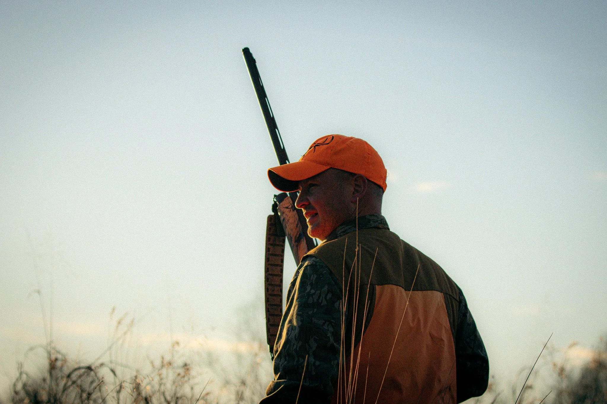 A person wearing an orange cap and a dark jacket holding a long firearm, with tall grass and a clear sky in the background.