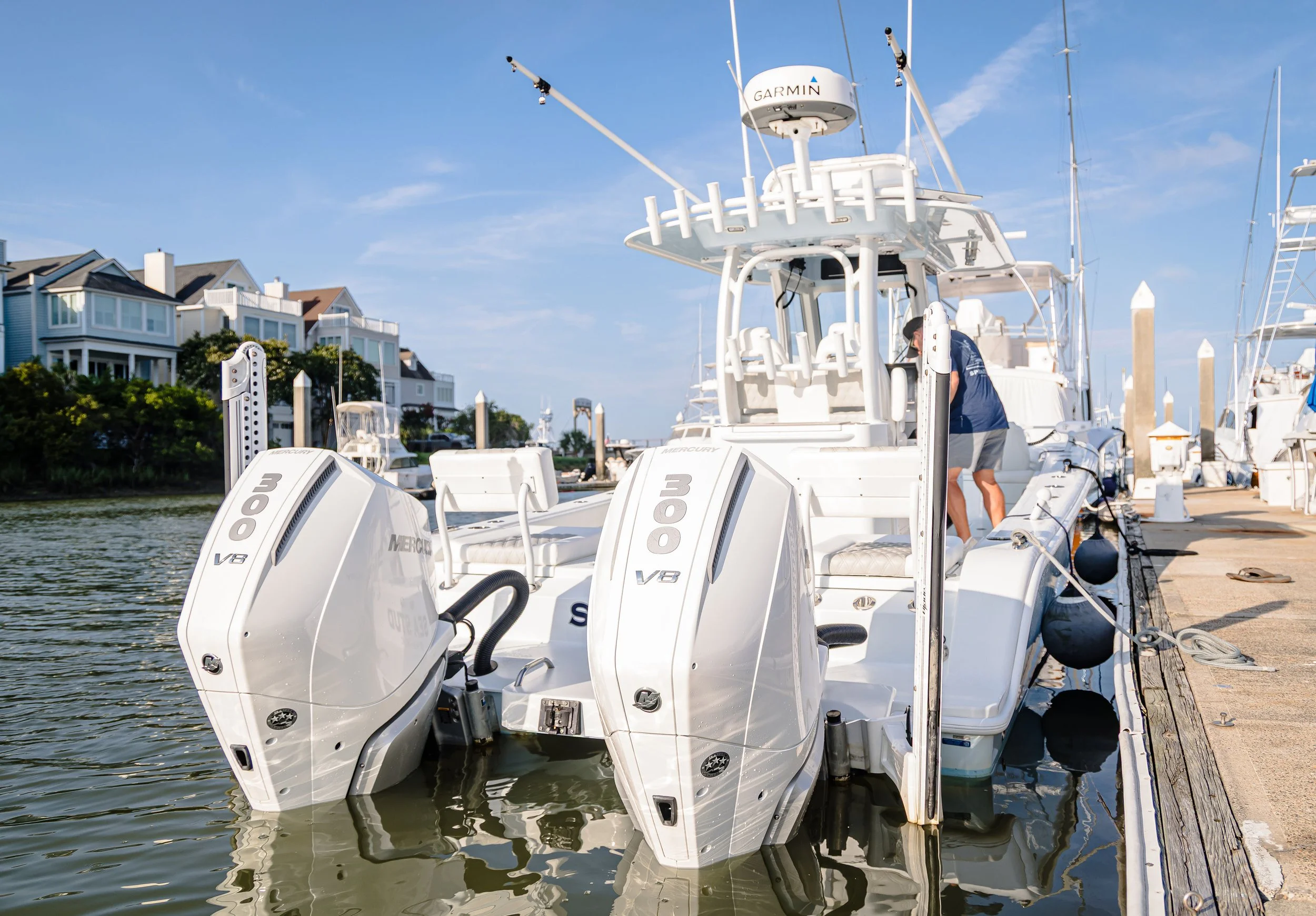 Close-up view of multiple white fishing boats with outboard motors docked at a marina, with residential houses and blue sky in the background.