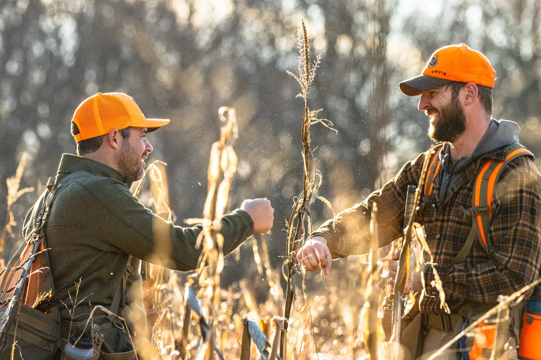 Two men in orange hunting hats and outdoor gear shaking hands in a wheat field during sunset.