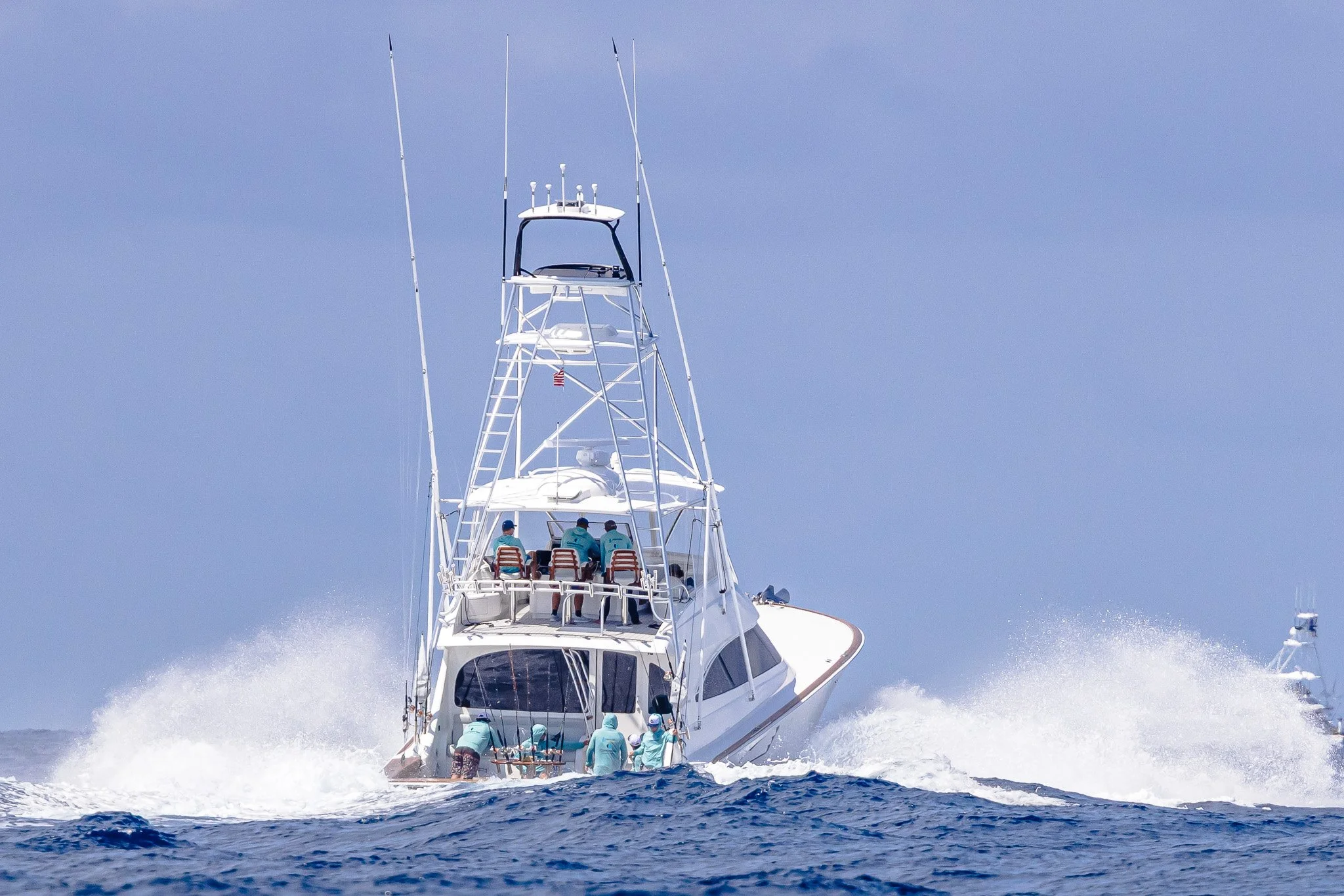 A large white yacht with multiple levels and antennas cruising on the open water, with several people on board wearing light blue shirts and hats. The yacht is moving quickly, creating waves and spray behind it, under a clear blue sky.