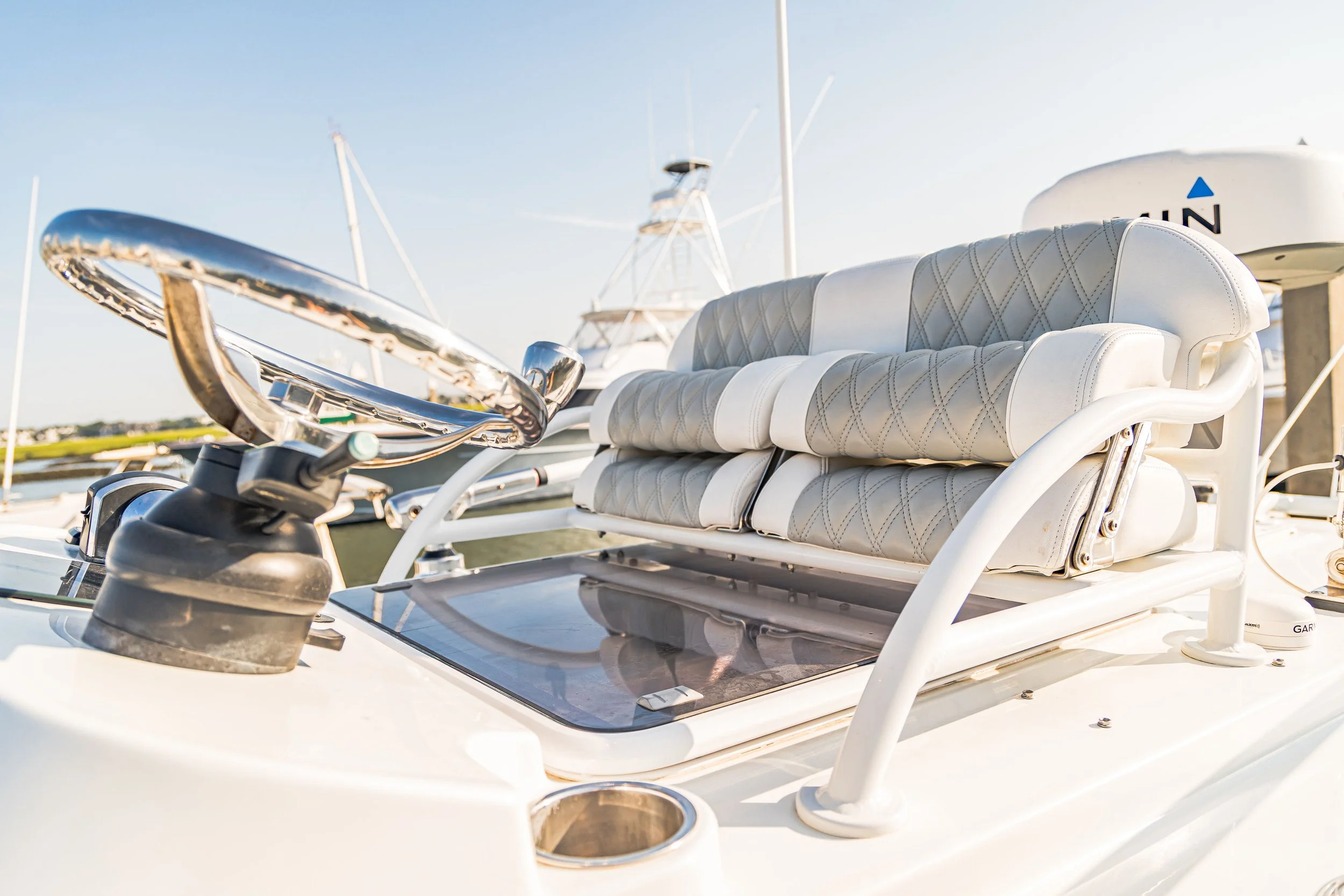 View of a white boat dashboard with a steering wheel, cushioned seating, and navigation equipment, docked at a marina with sailboats in the background.