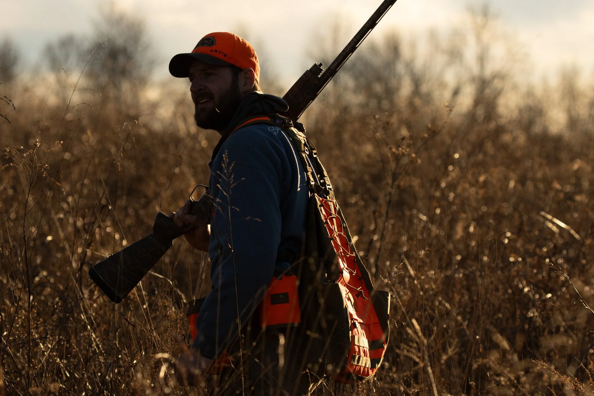 A man outdoors in a field during sunset, wearing camouflage clothing, an orange cap, and carrying a rifle on his shoulder. He appears to be hunting.