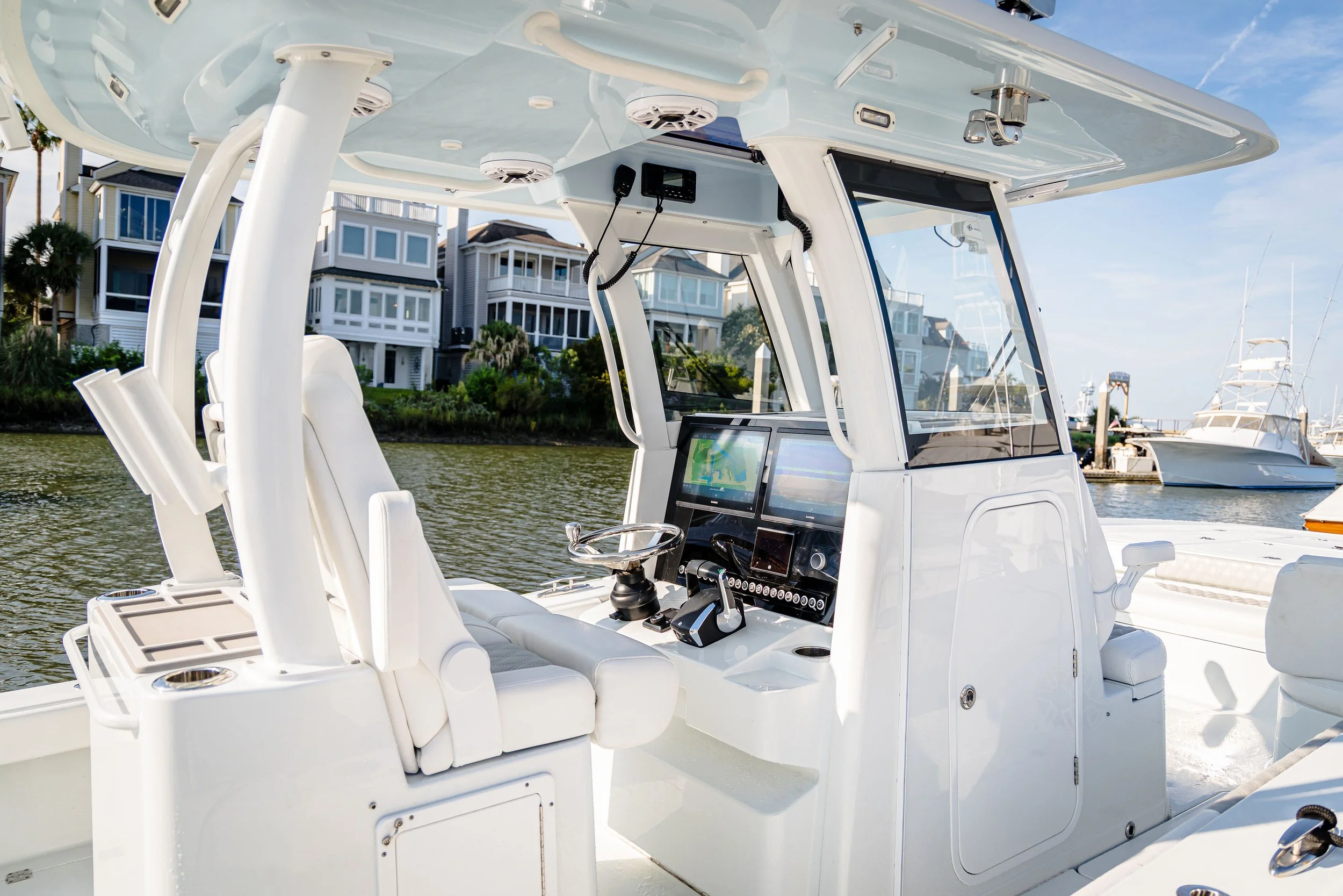 The interior of a white boat with steering controls, navigation screens, and seating, docked on a waterway with houses and other boats in the background under a clear blue sky.