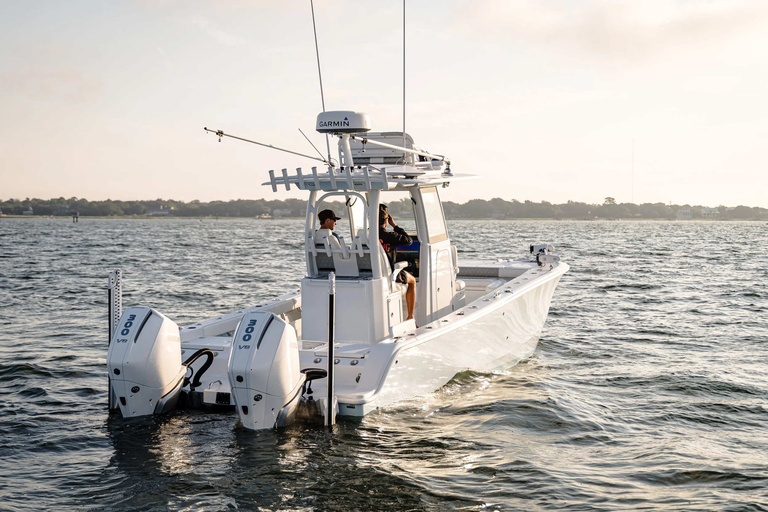A white fishing or chase boat with two outboard engines moving on the water during sunset, with two people onboard, one sitting and the other standing, looking out over the water.